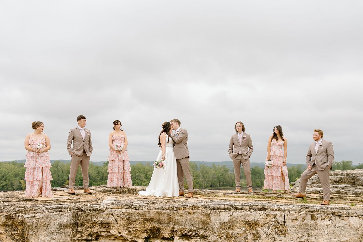 Wedding party stands on stone wall against cloudy sky with bridesmaids in pink and groomsmen in tan suits.