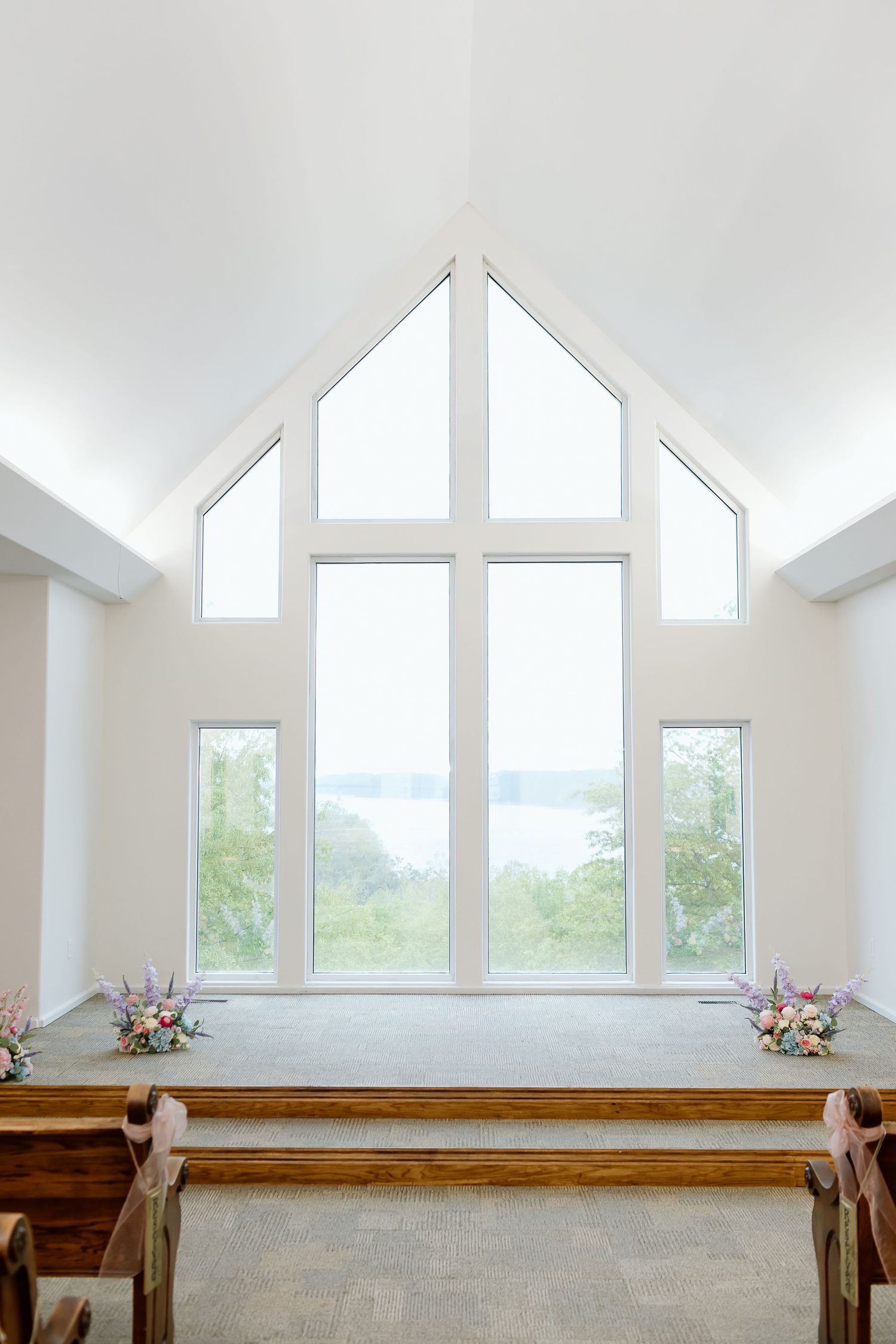 Modern chapel interior with large triangular window, wooden pews and floral arrangements overlooking a scenic lake view.