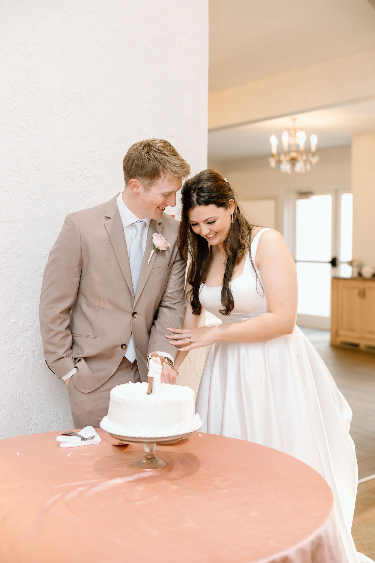 A couple in wedding attire shares a sweet moment while cutting their wedding cake in a bright indoor venue.