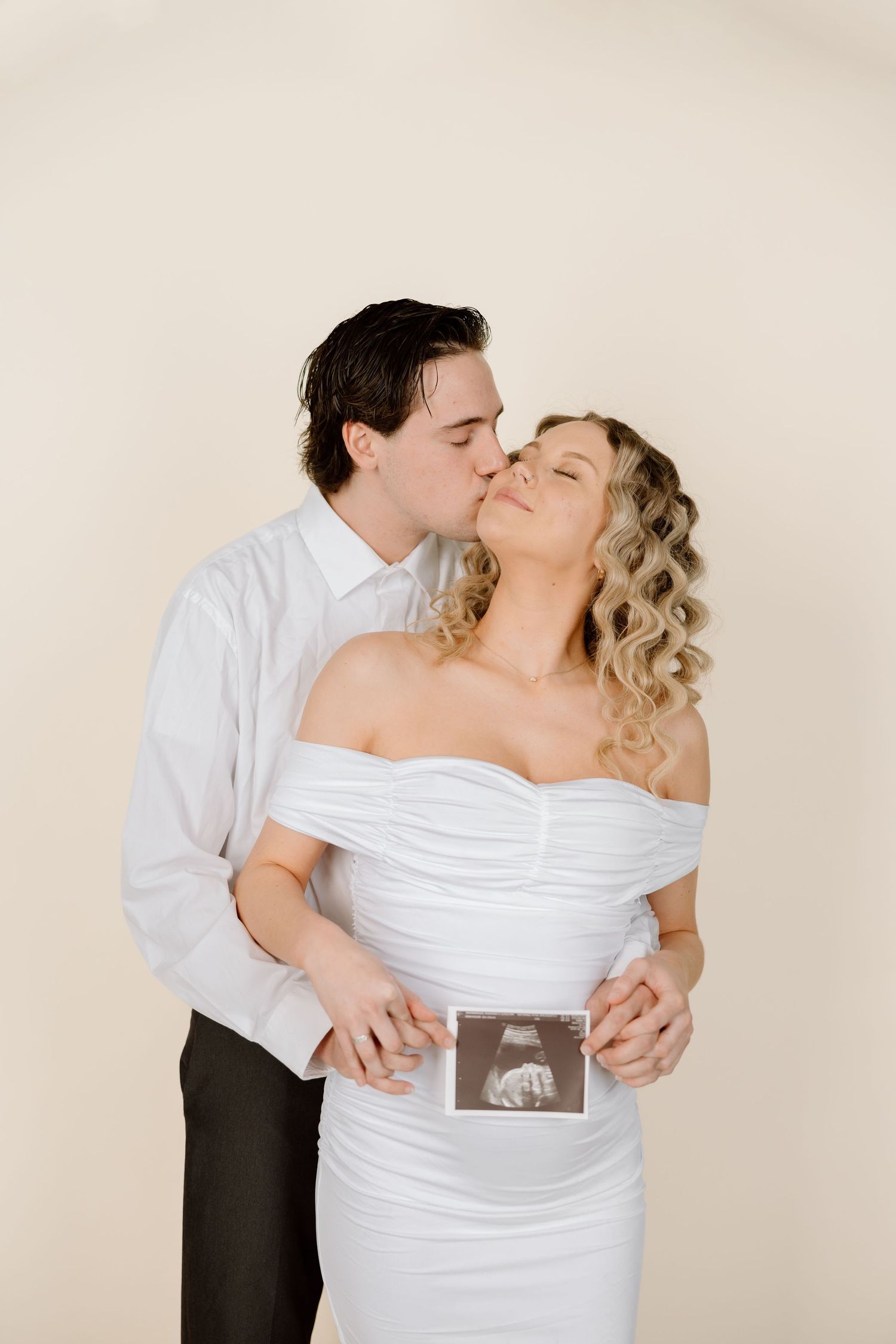 Romantic couple in white and black attire share tender moments while holding ultrasound photo in maternity shoot.