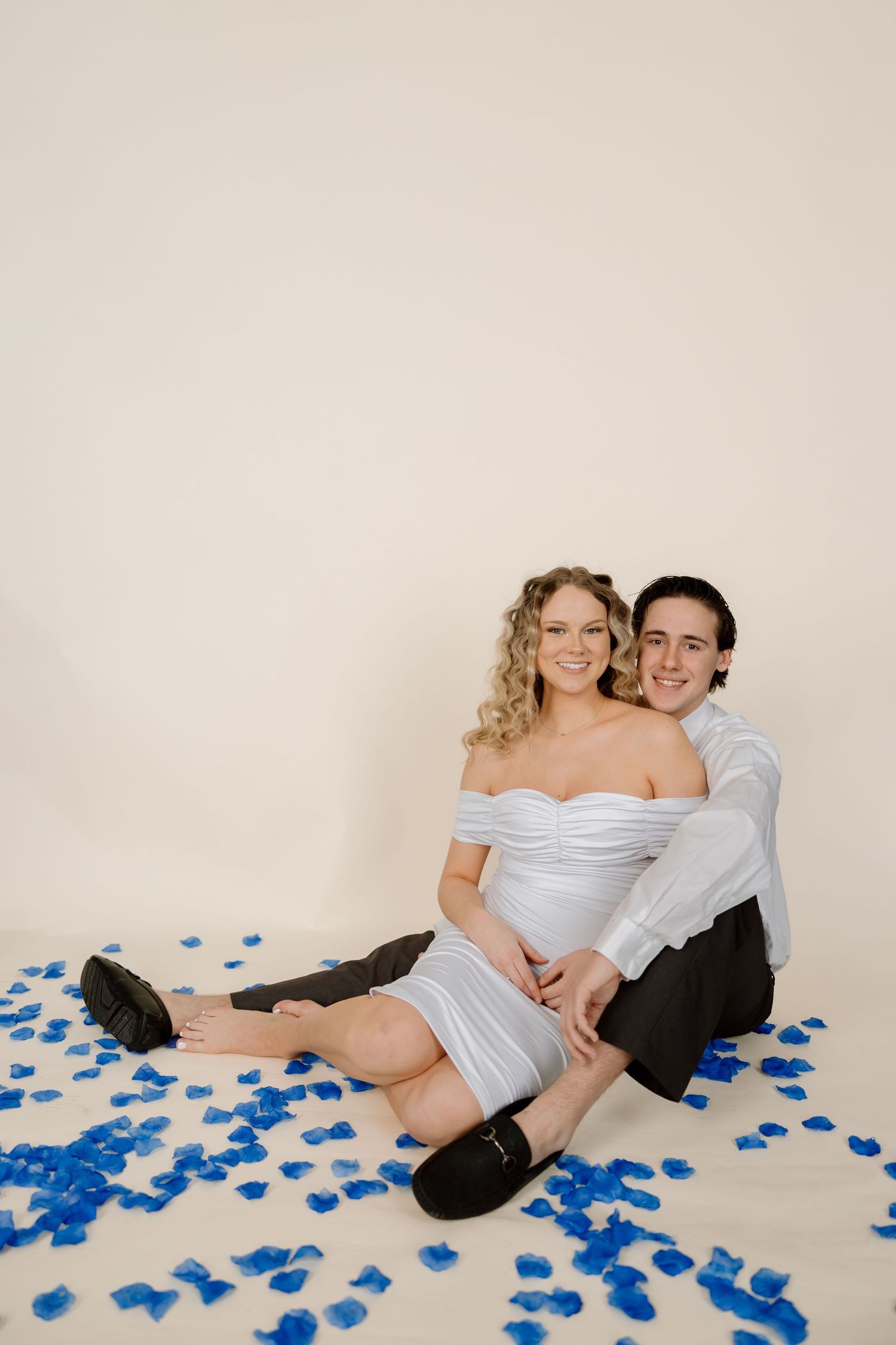 Couple in formal attire poses intimately on floor with scattered blue rose petals in romantic photoshoot series.