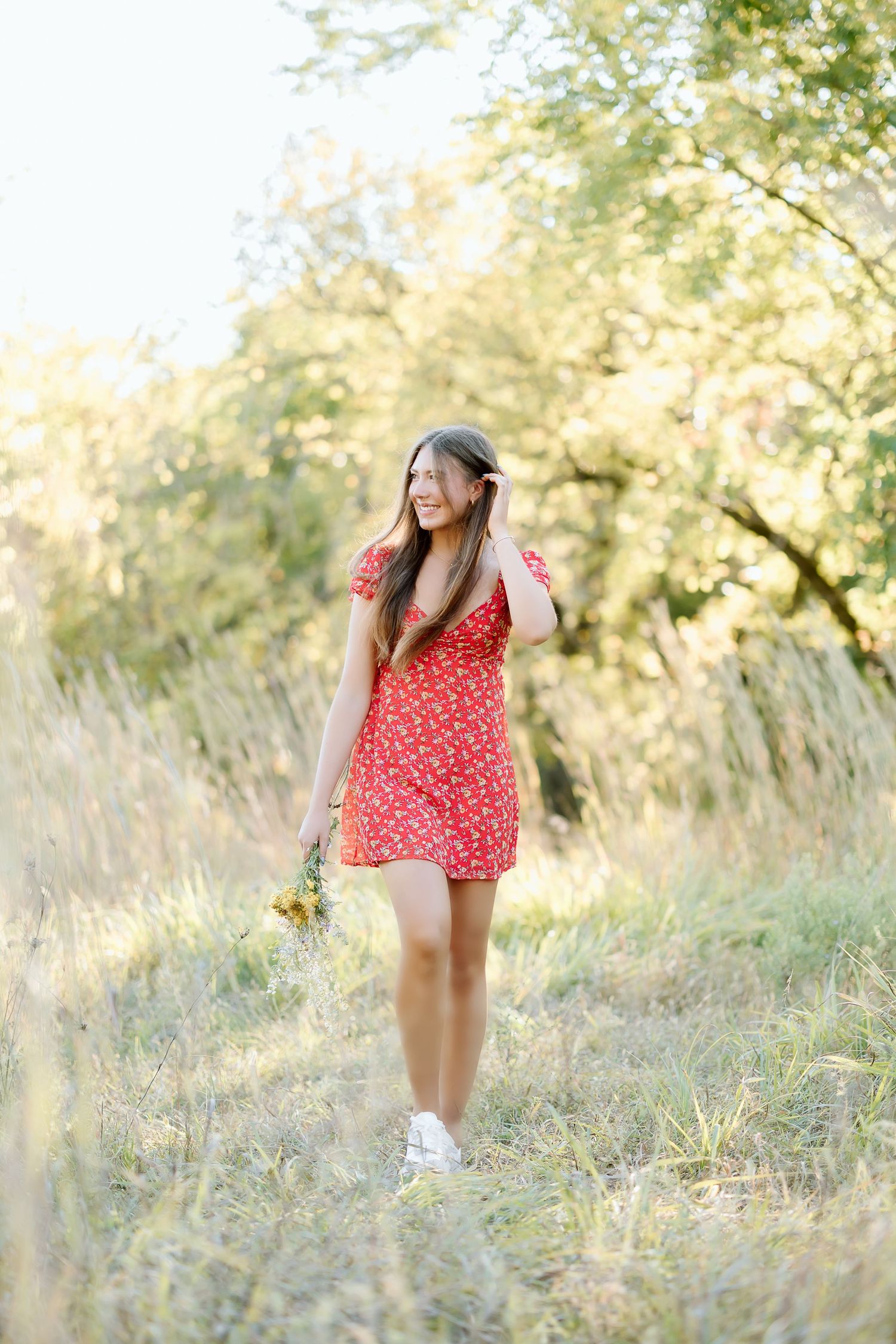 A person in a red floral dress stands in a sunlit meadow with tall grass and trees in the background.