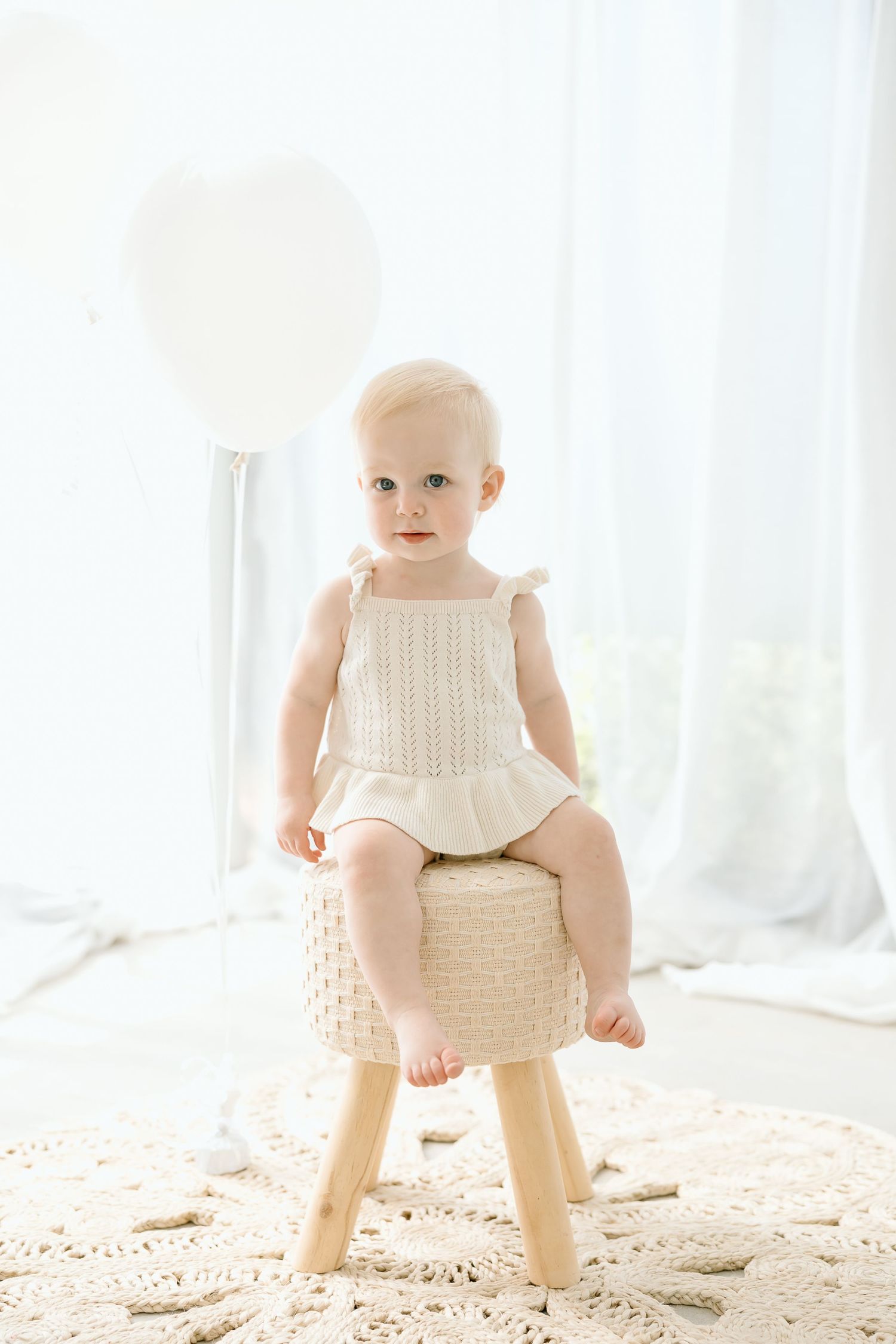 A baby in a cream knit outfit sits on a small wooden stool in a bright, sunlit room with white curtains.