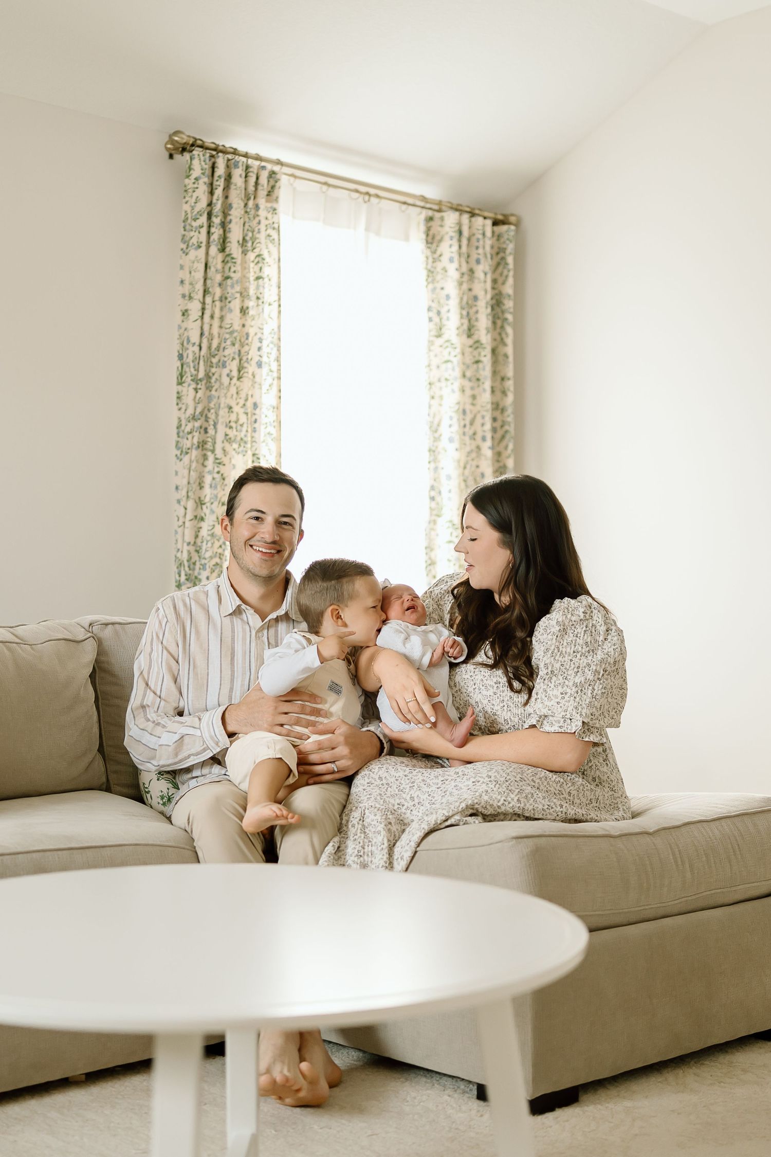 Family relaxes together on beige couch in bright living room with floral curtains.