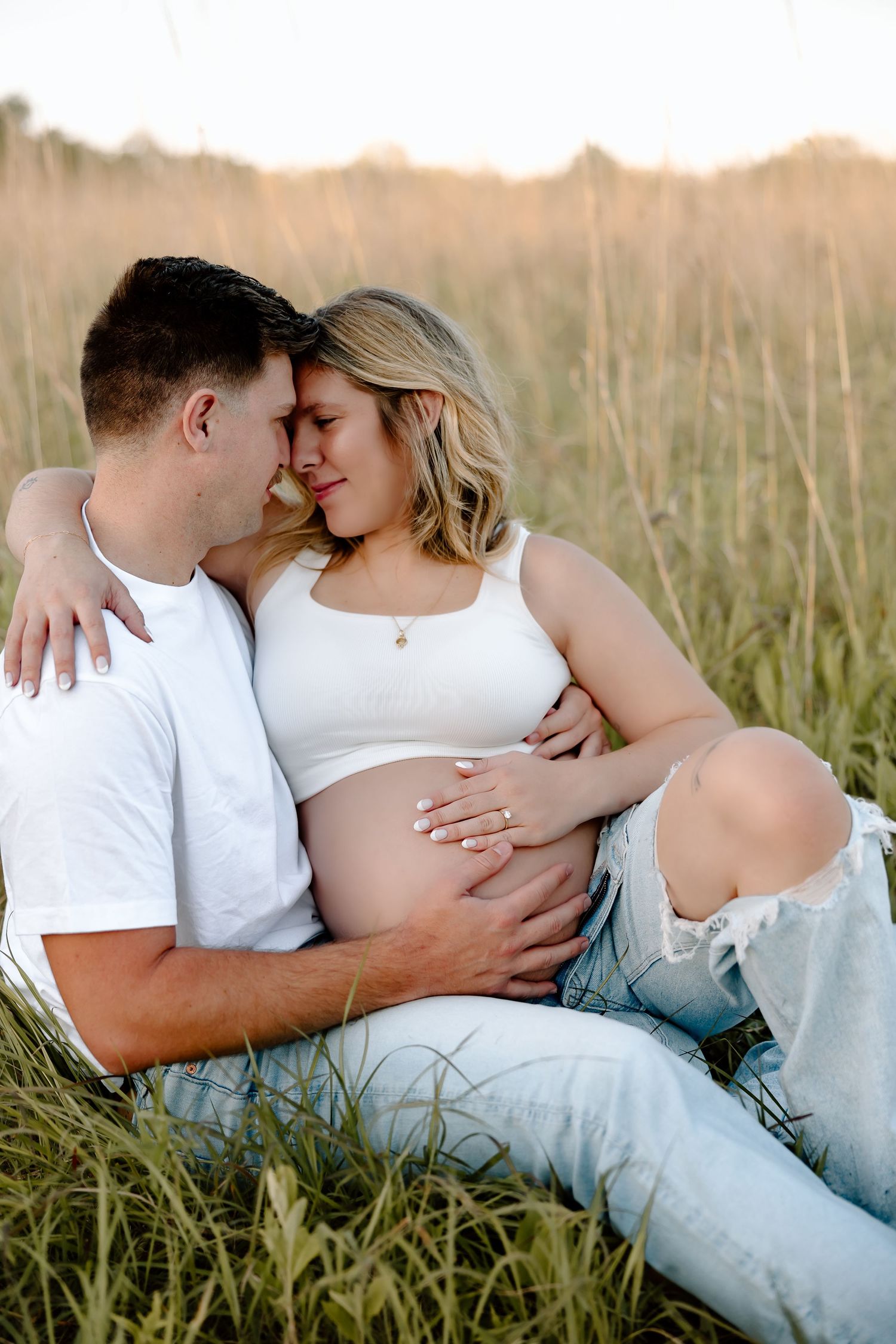 A couple in white shirts and jeans embrace intimately in a golden sunlit field of tall grass at sunset.
