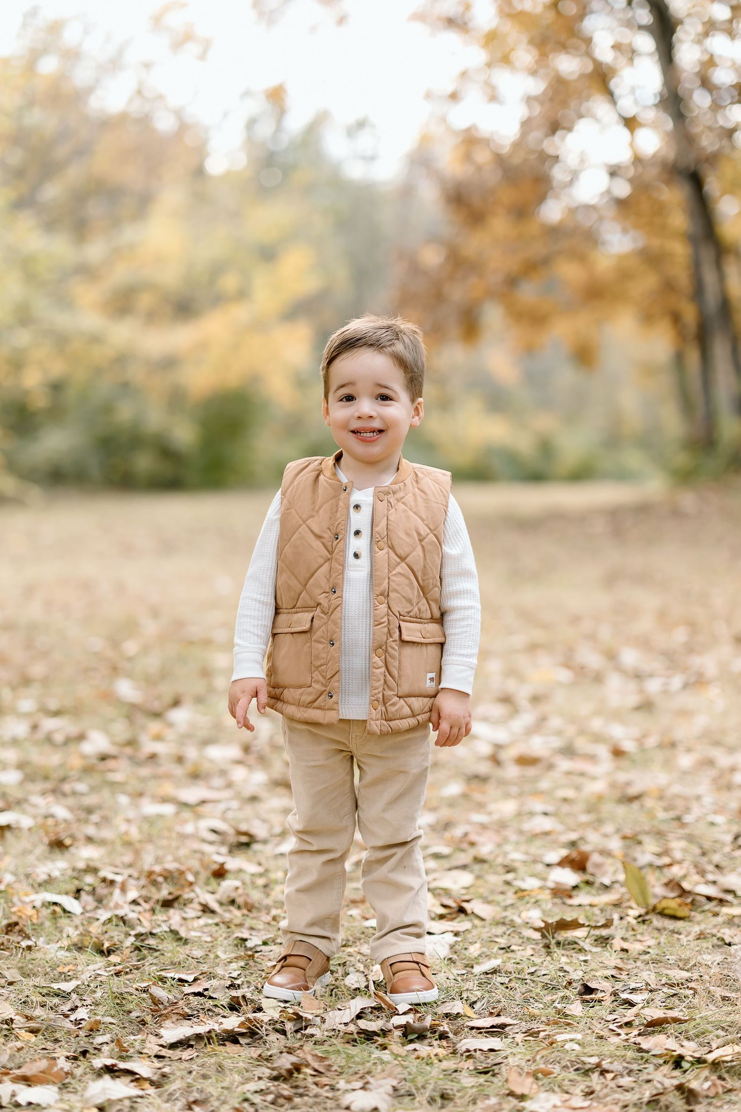 Small child wearing tan quilted vest and khaki pants stands smiling among fallen autumn leaves in a park setting.