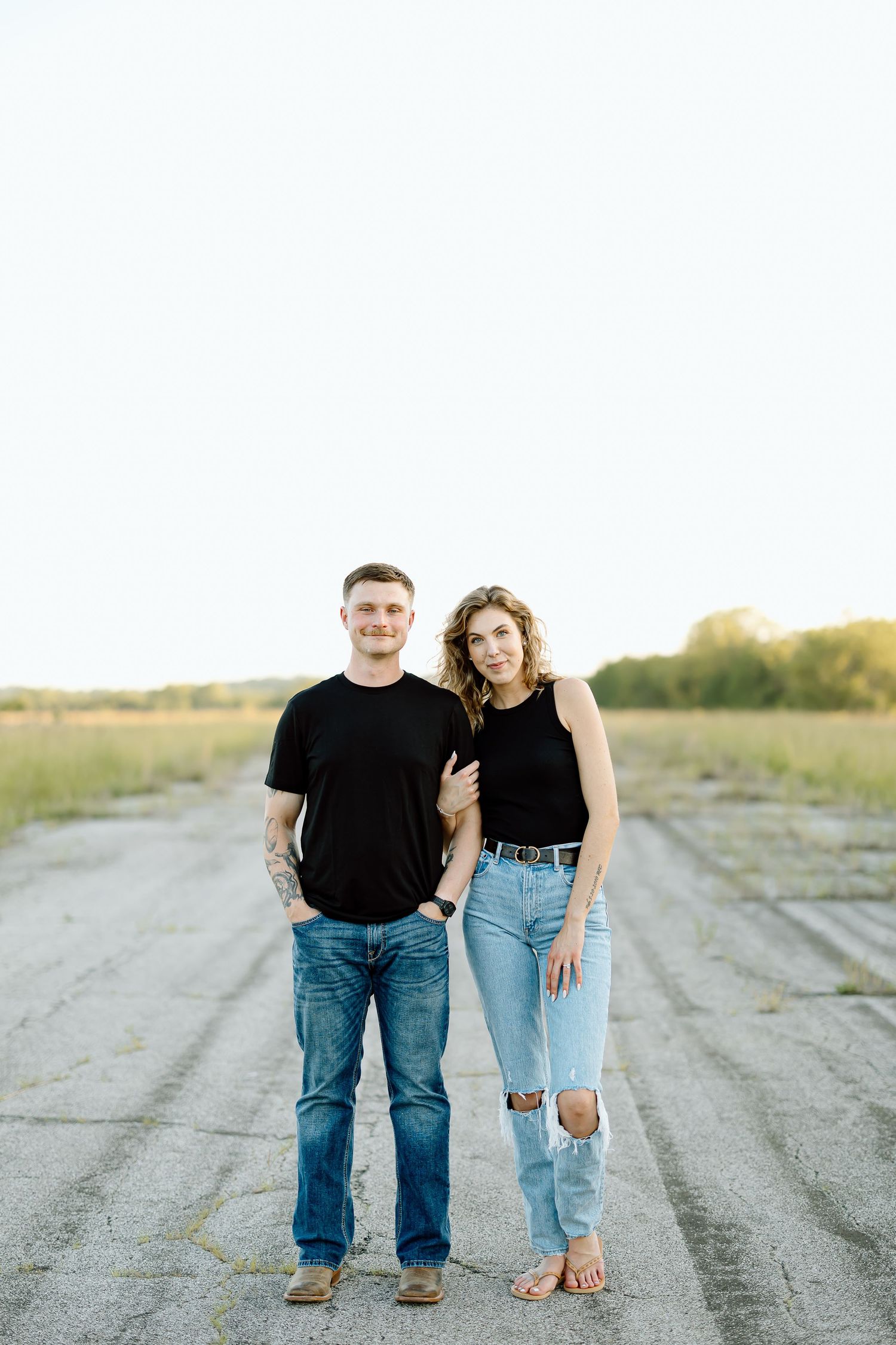 A couple in matching black tops and blue jeans stands together on a dirt road at sunset.