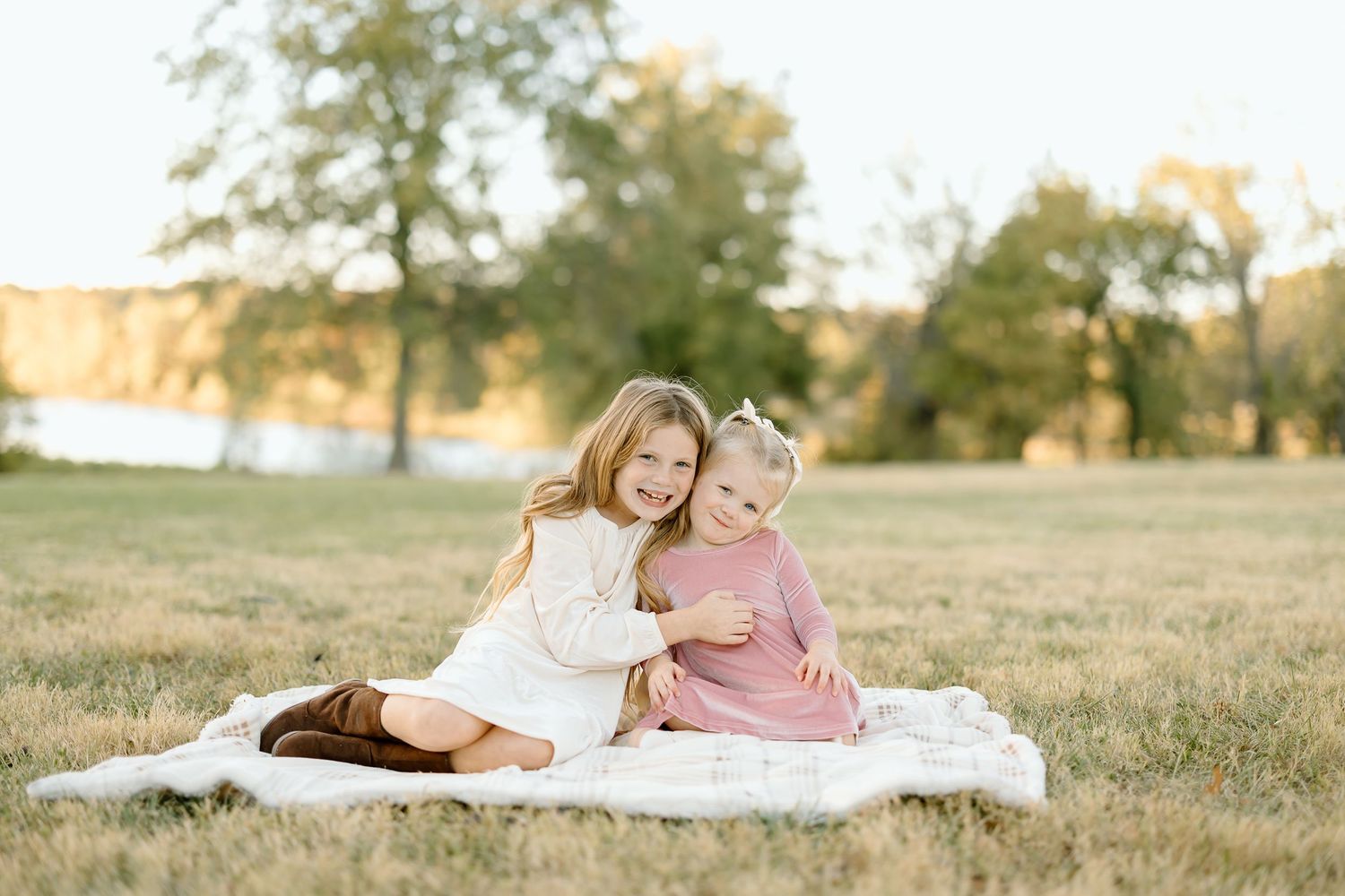 Two people in coordinating white and pink outfits sit together on a blanket in a sunlit grassy field.