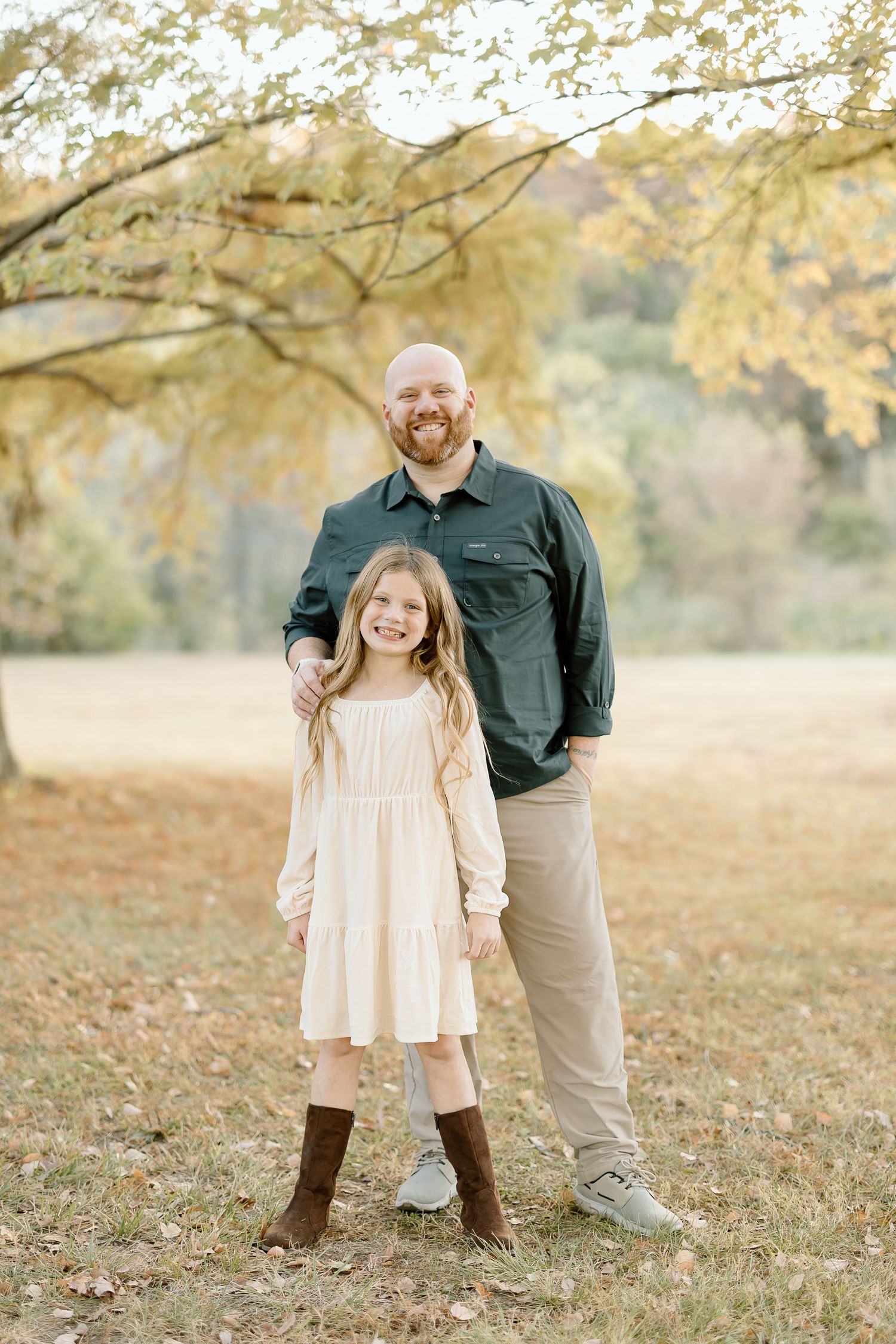 A parent and child stand together under golden autumn trees in a field wearing casual clothing and boots.