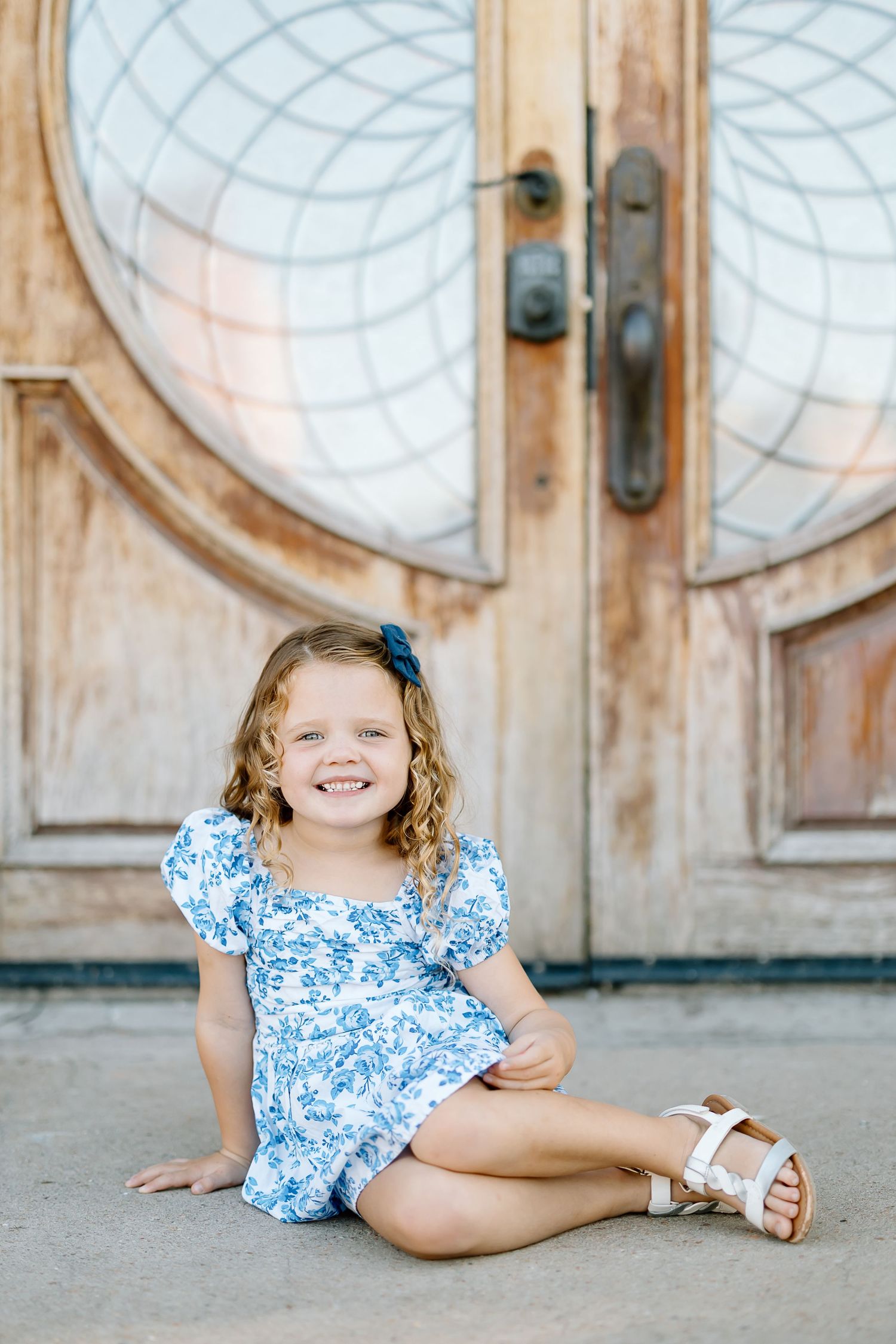 A child in a blue and white floral dress poses playfully on a concrete floor in front of rustic wooden doors.
