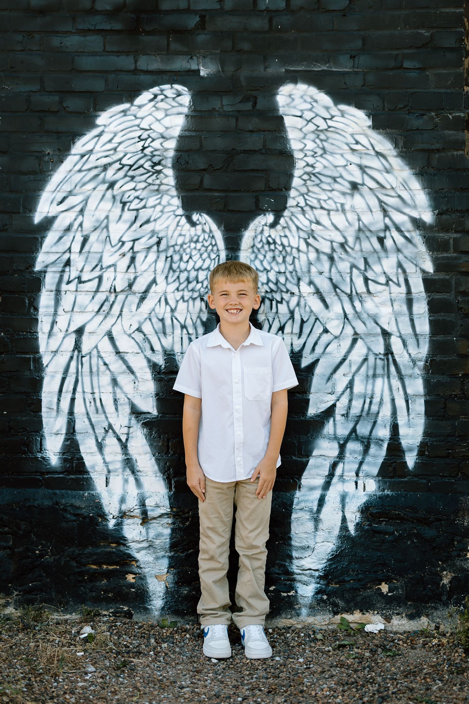 A child stands in front of a white angel wings mural painted on a dark brick wall.