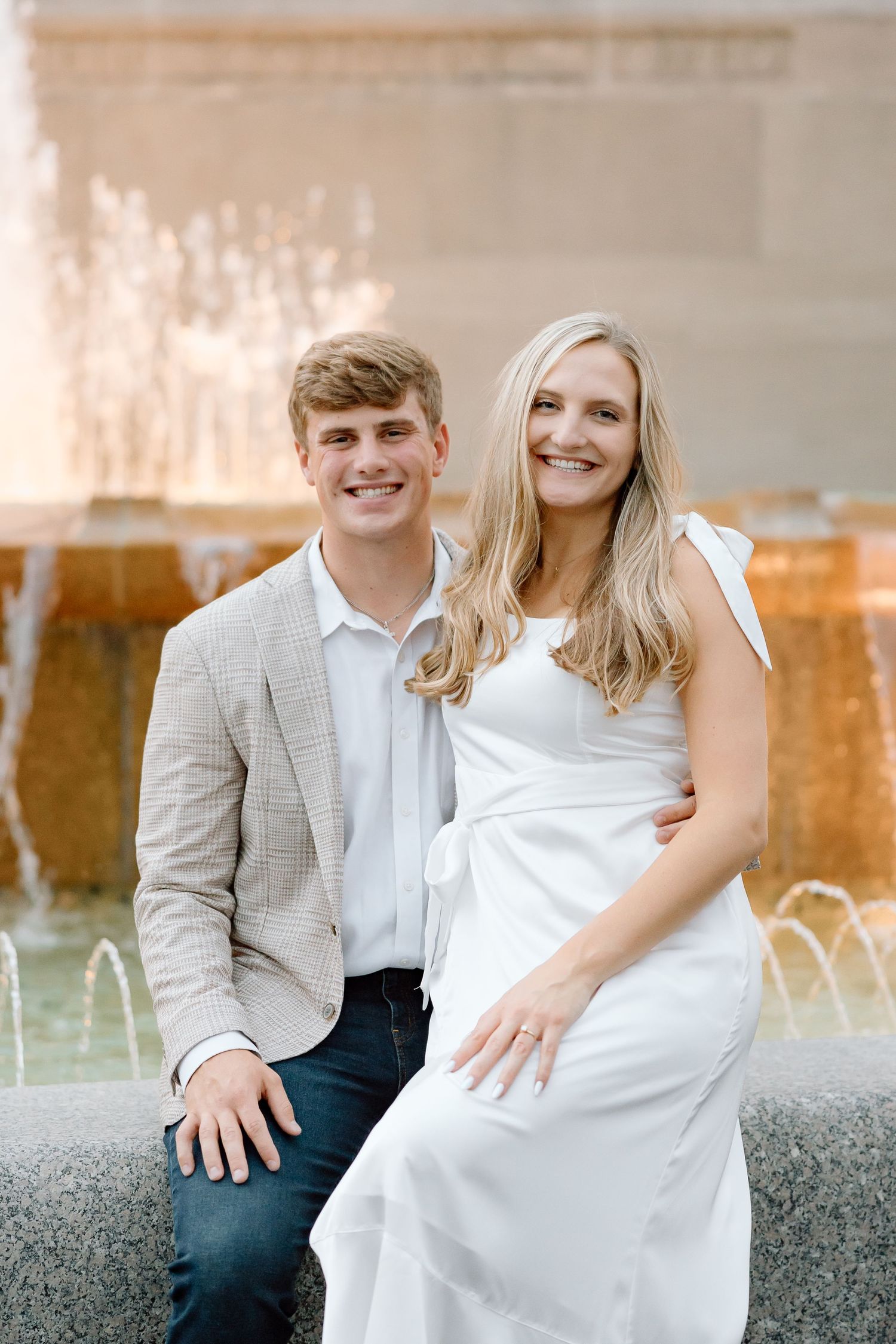 A couple sits together on a concrete ledge during a romantic outdoor engagement photo session at sunset.