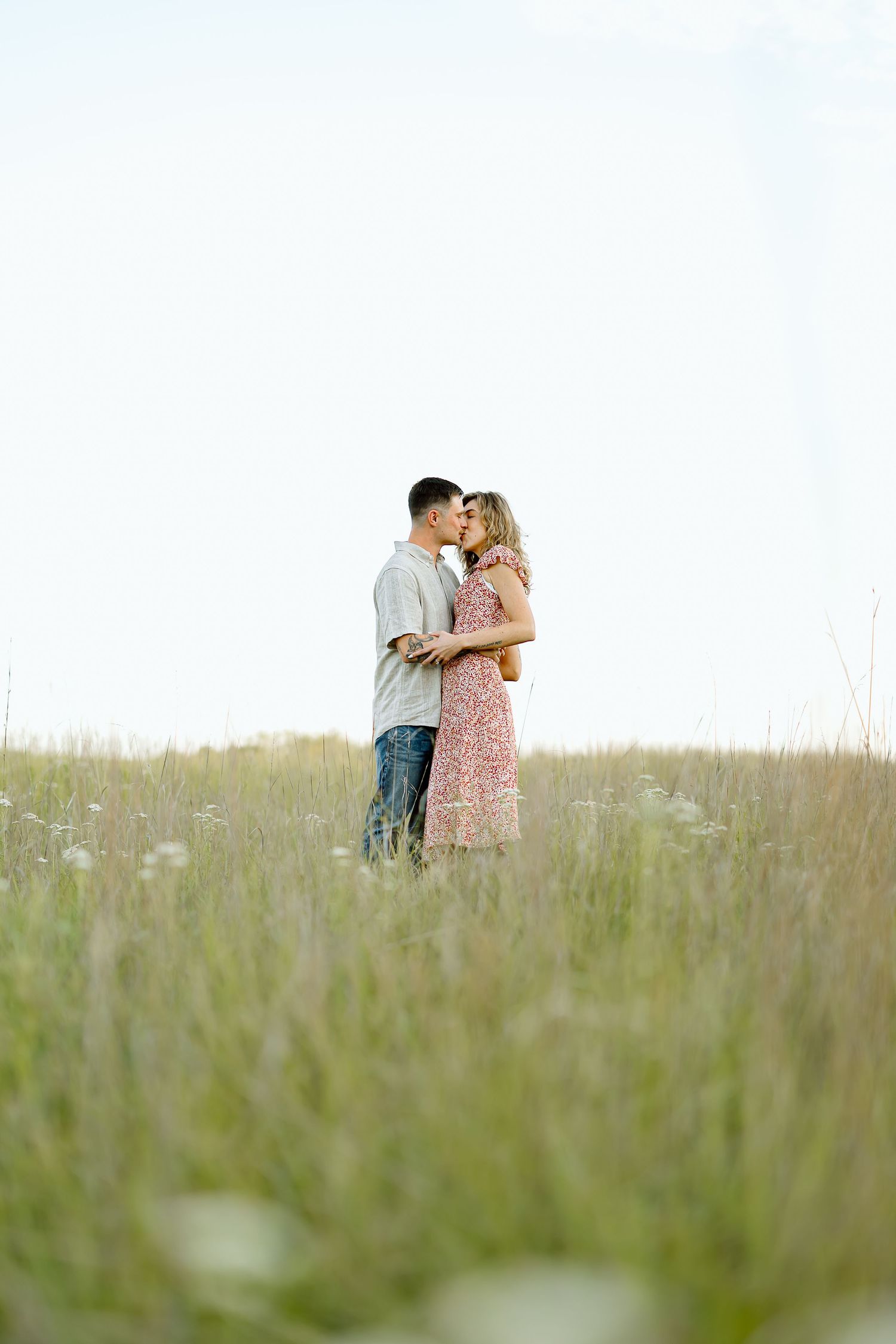A couple shares a tender moment while standing in a tall grass meadow on a bright summer day.