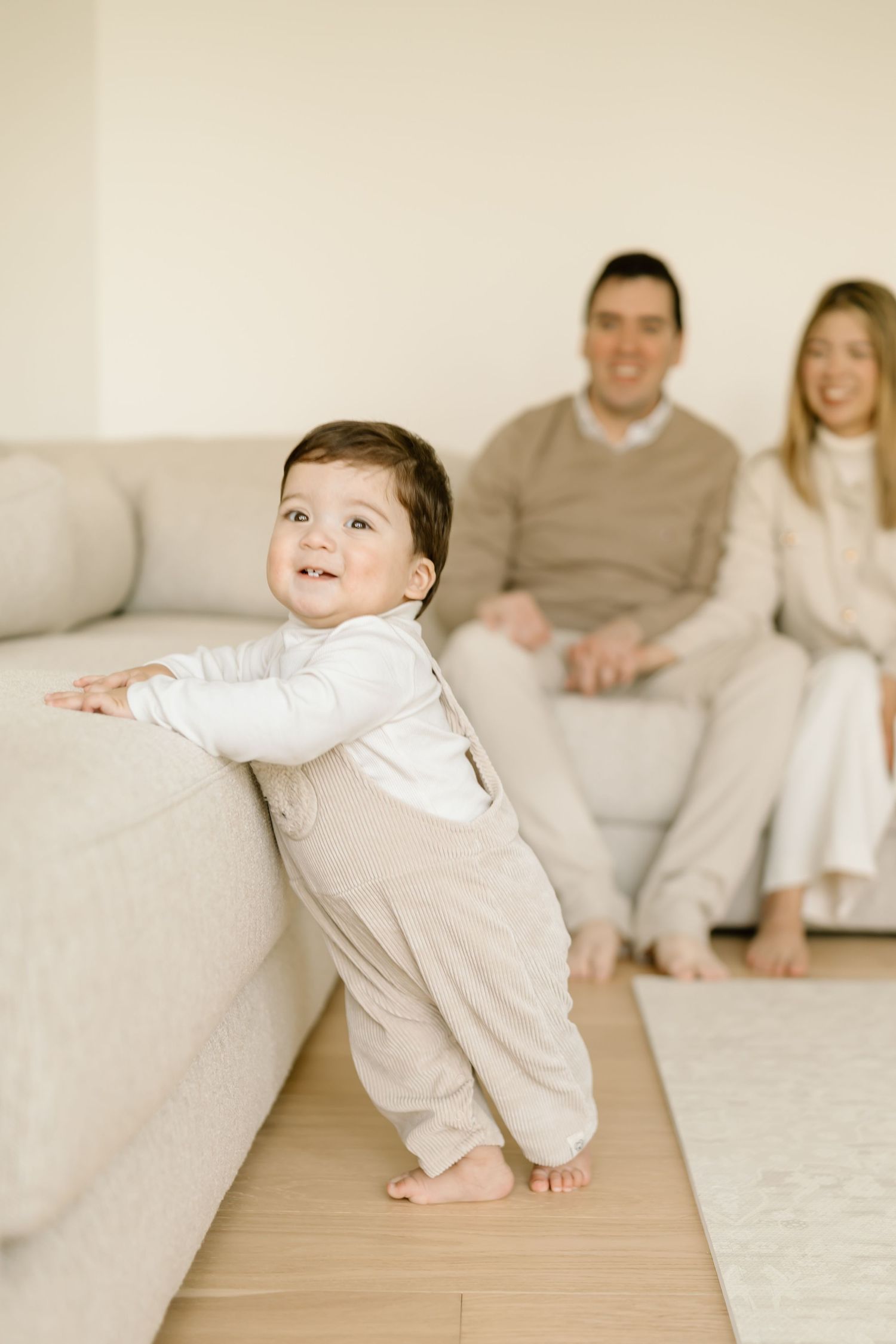 Baby in white onesie crawls on wooden floor while family sits on couch in background.