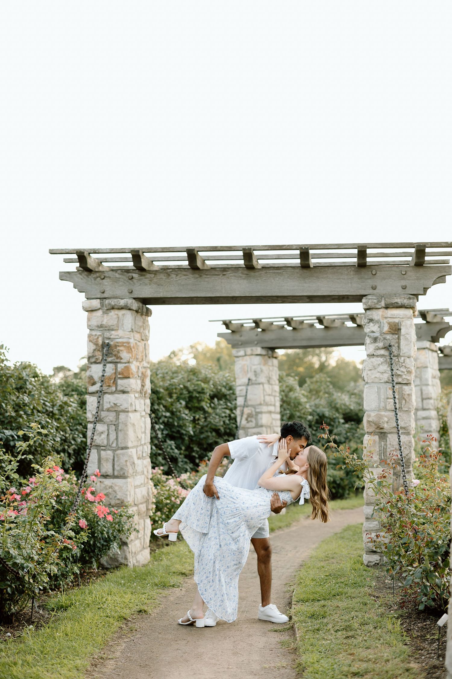Couple shares romantic moments under rustic stone pergola surrounded by blooming rose garden.
