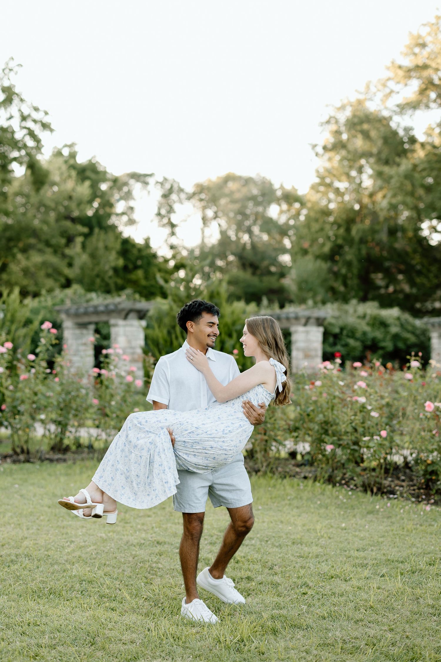 A couple in white outfits dances joyfully together in a garden setting during golden hour.