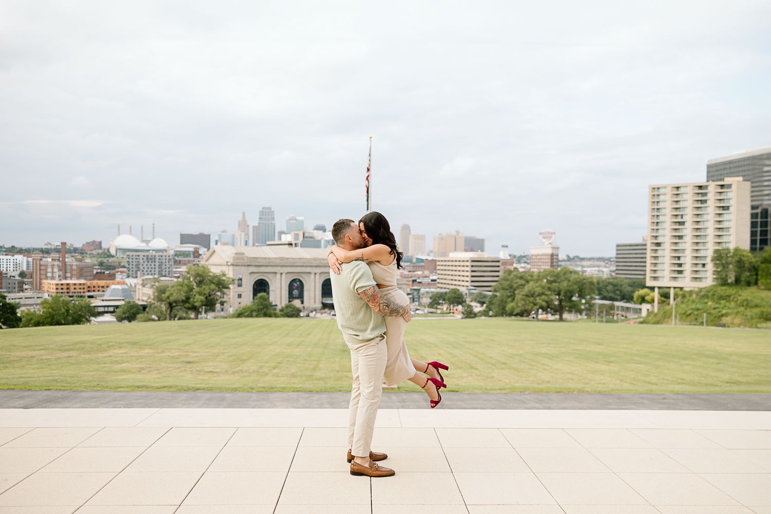 Romantic couple embraces in a scenic outdoor location with city buildings and greenery in the background.