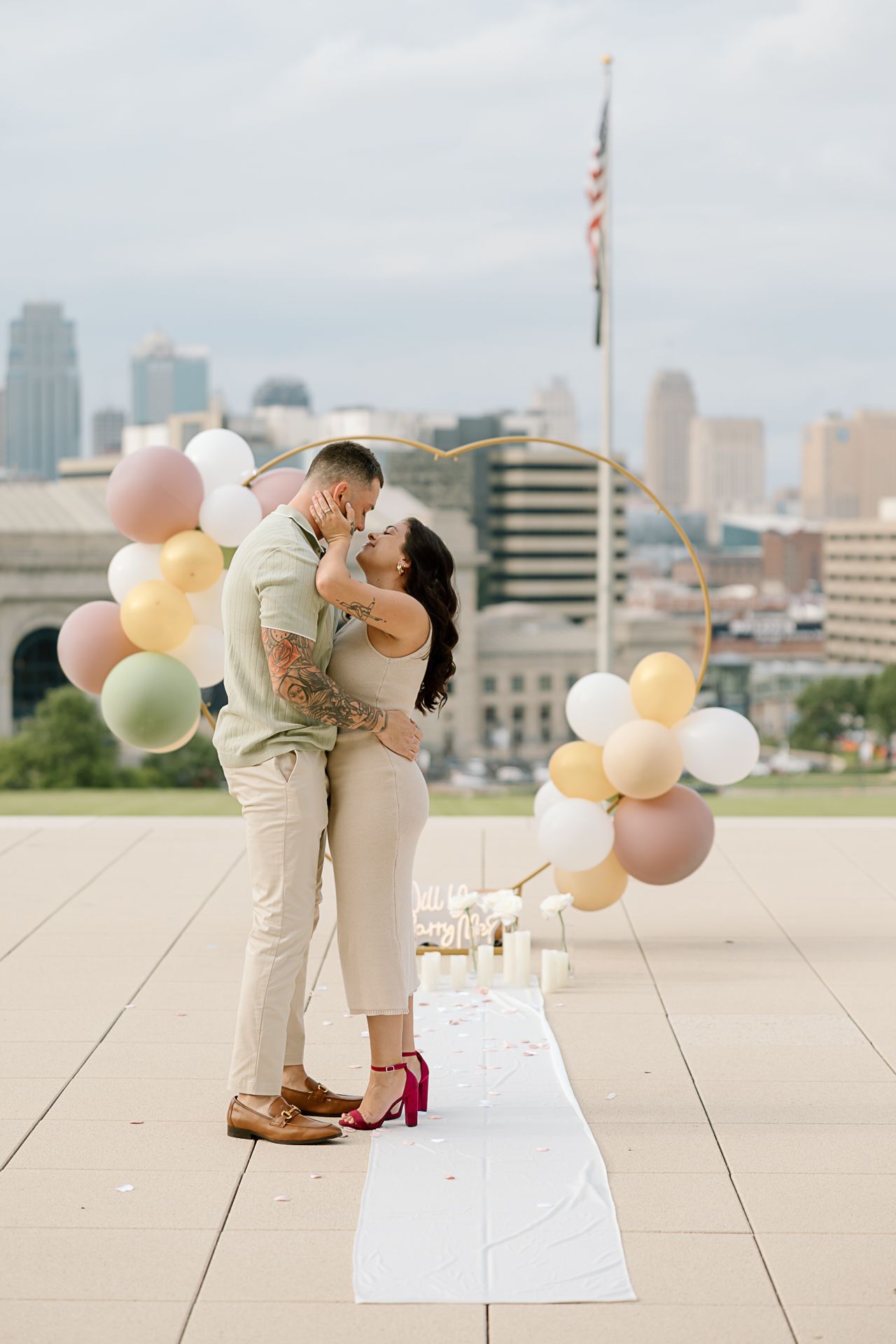 A couple shares a celebratory embrace during a rooftop proposal with colorful balloons and urban skyline views.