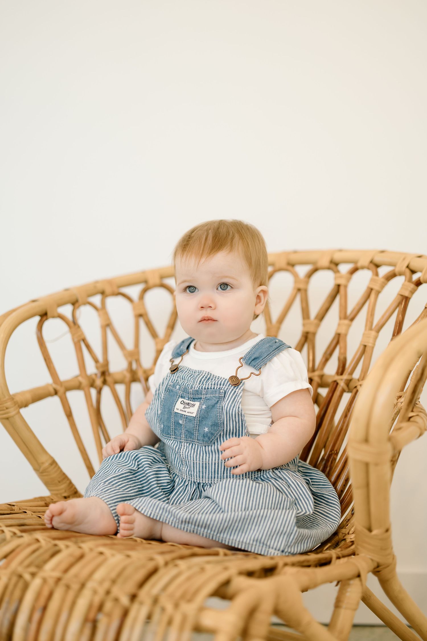 A small child in a blue striped dress sits on a curved rattan bamboo chair against a white background.