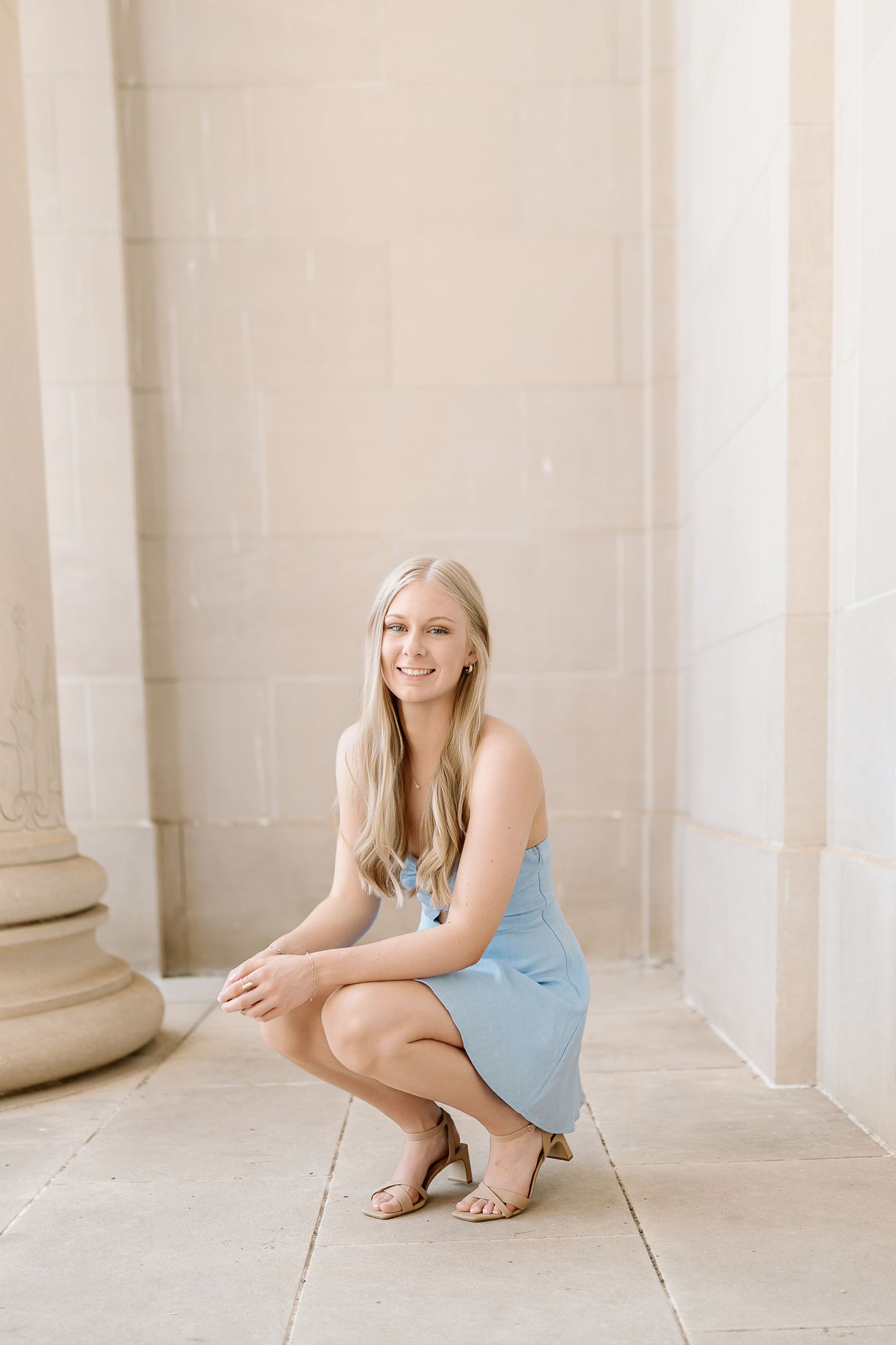 A person in a blue dress poses elegantly against white columns in a classic architectural setting.