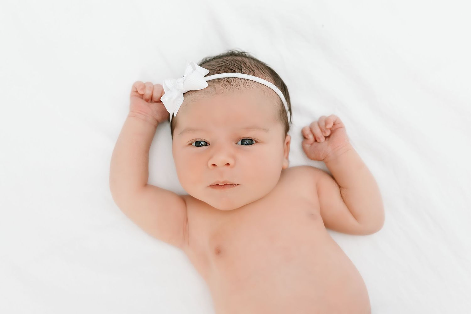Sweet baby in white headband lying on soft white blanket stretches arms while looking up at camera.