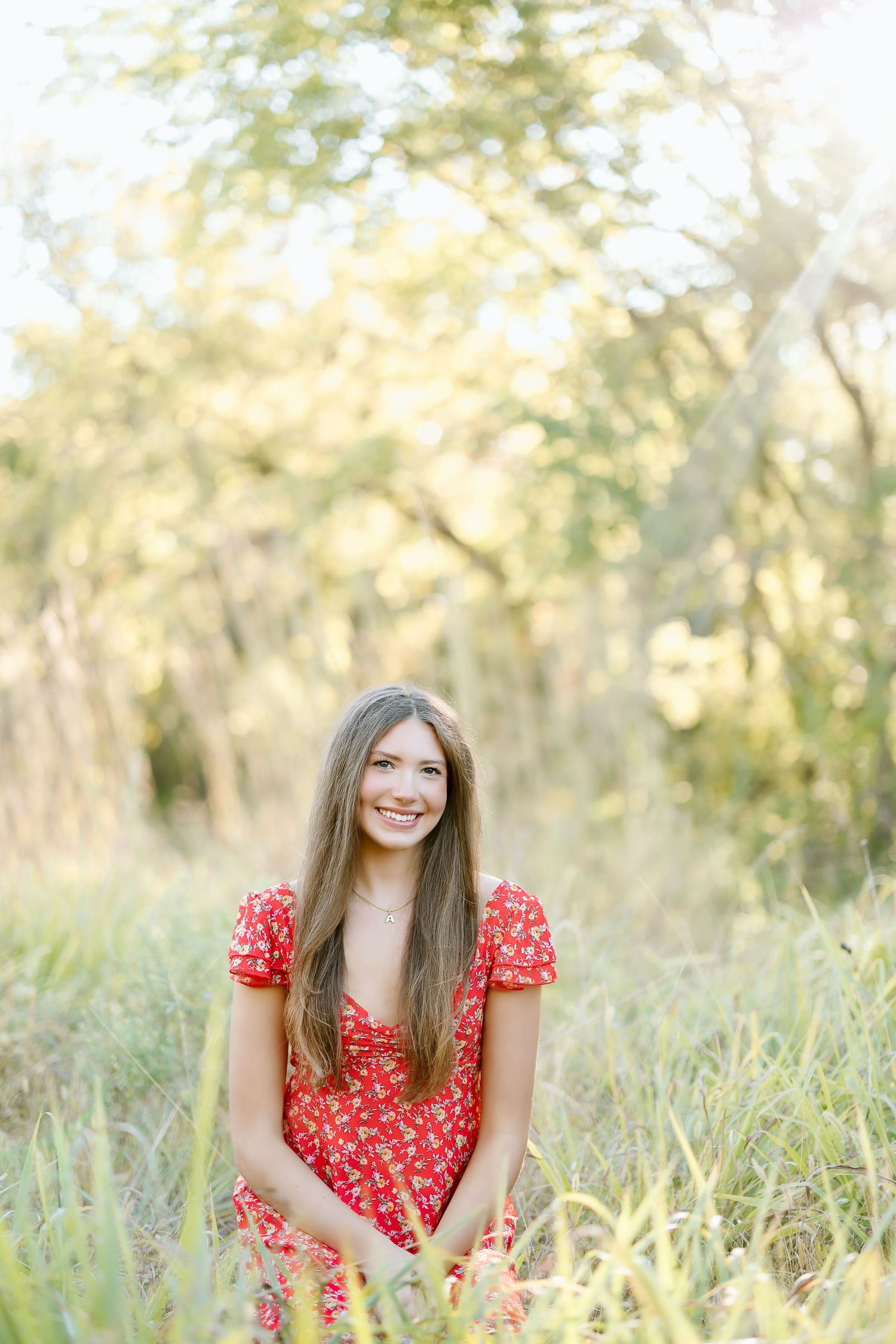 A person in a red floral dress sits in a sunny meadow with tall grass and trees in the soft golden light.