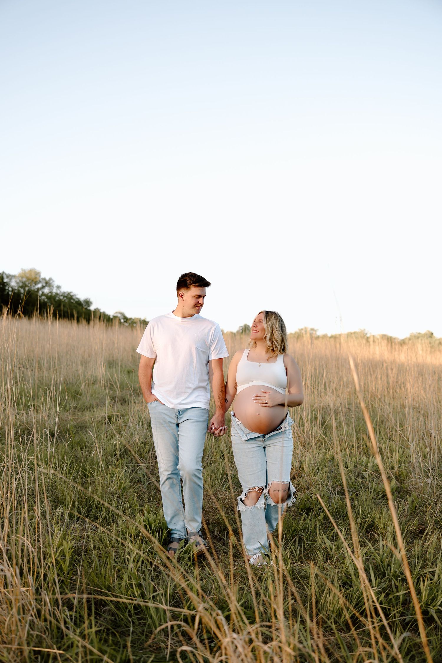 Couple holding hands while walking through a golden field of tall grass at sunset.