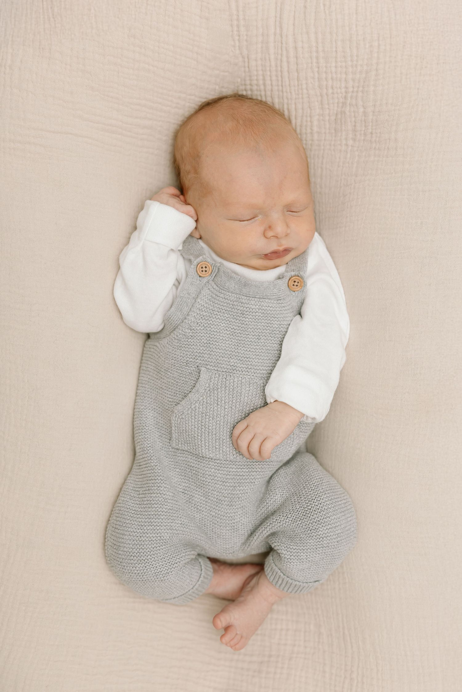Sleeping newborn baby wearing grey knit overalls and white shirt lies peacefully on cream textured blanket.