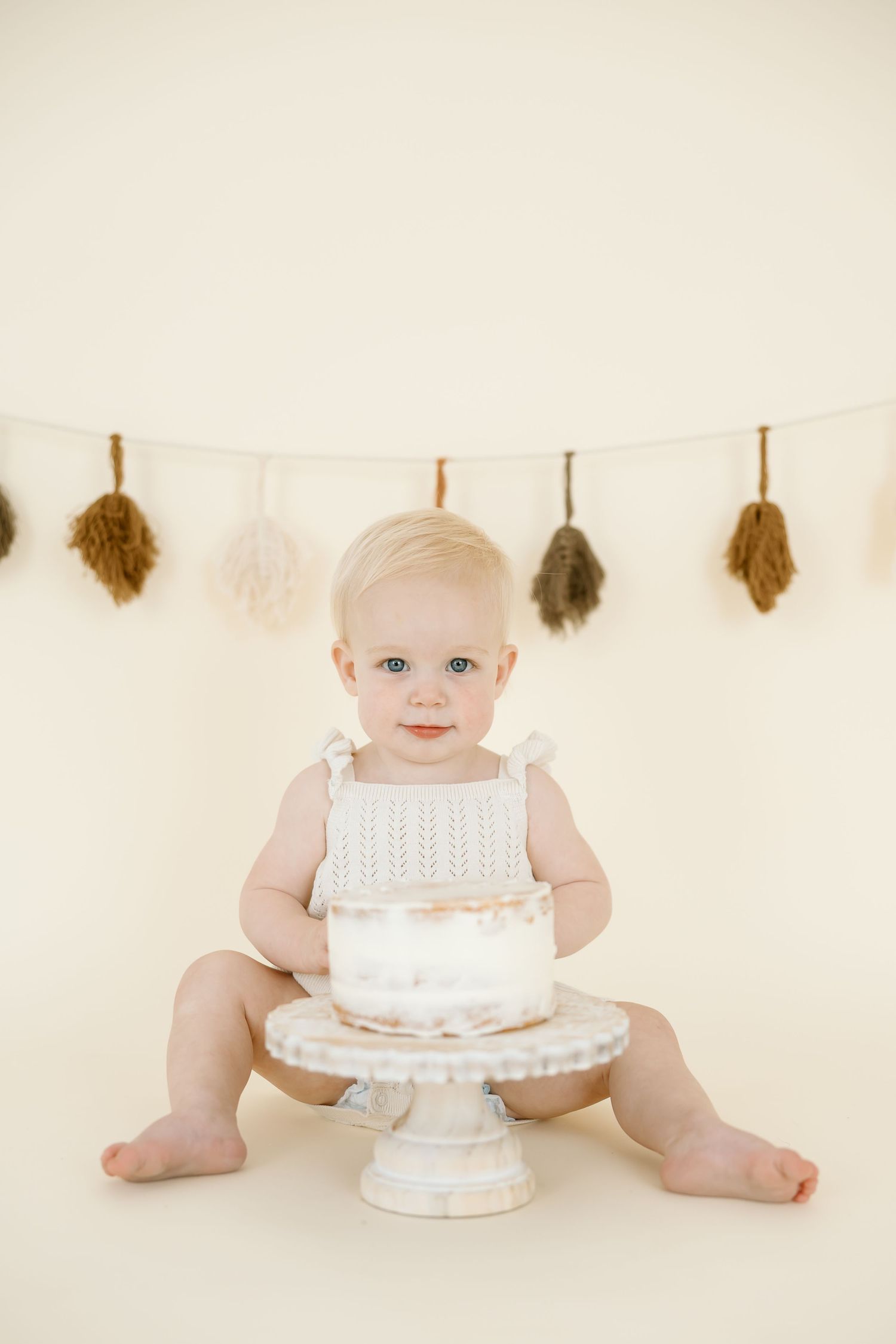 Baby in white eyelet dress sits with bare feet beside a white frosted cake during a first birthday photo session.