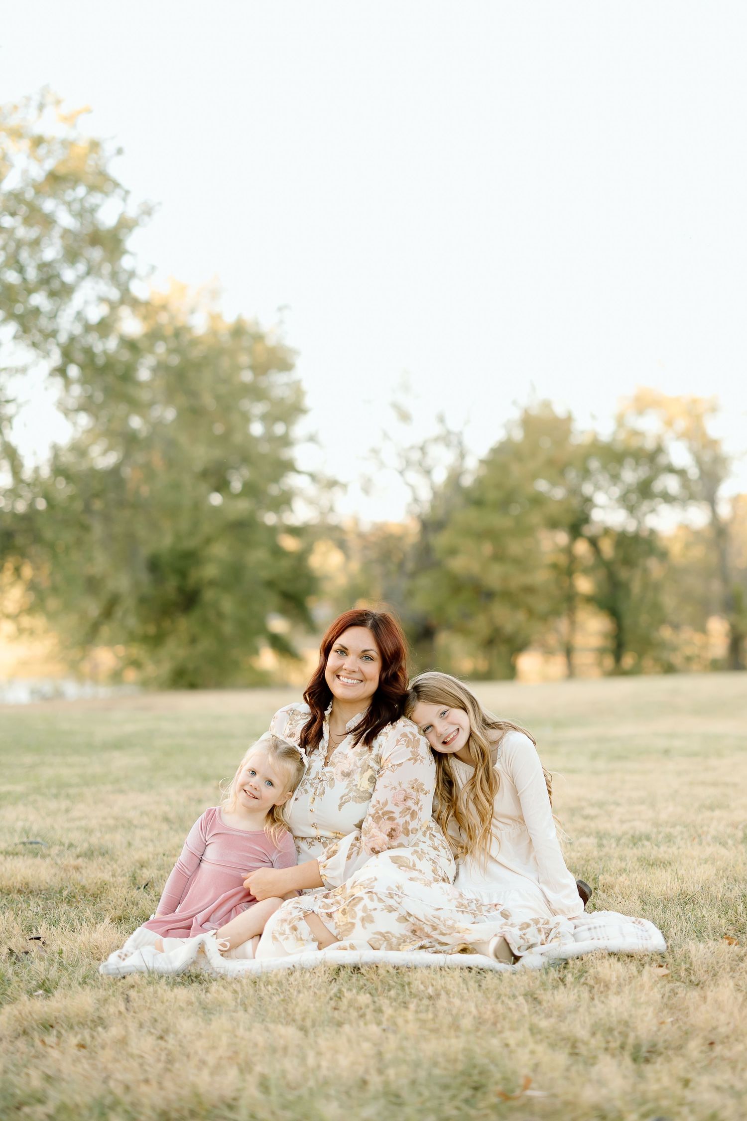 Three people laying in a grassy field at sunset wearing white and floral dresses.