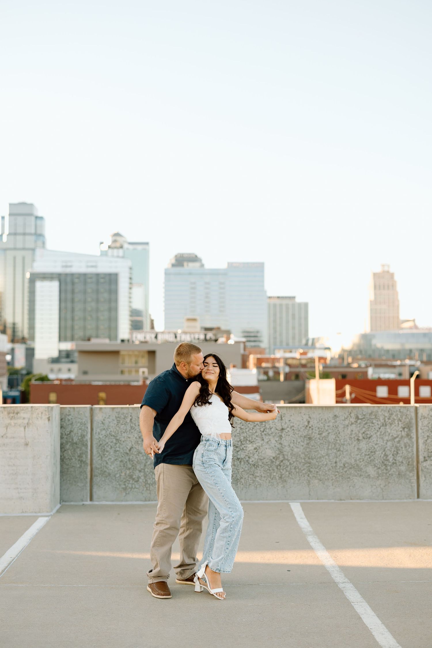 A couple embraces on a rooftop parking garage with a city skyline visible in the background at sunset.