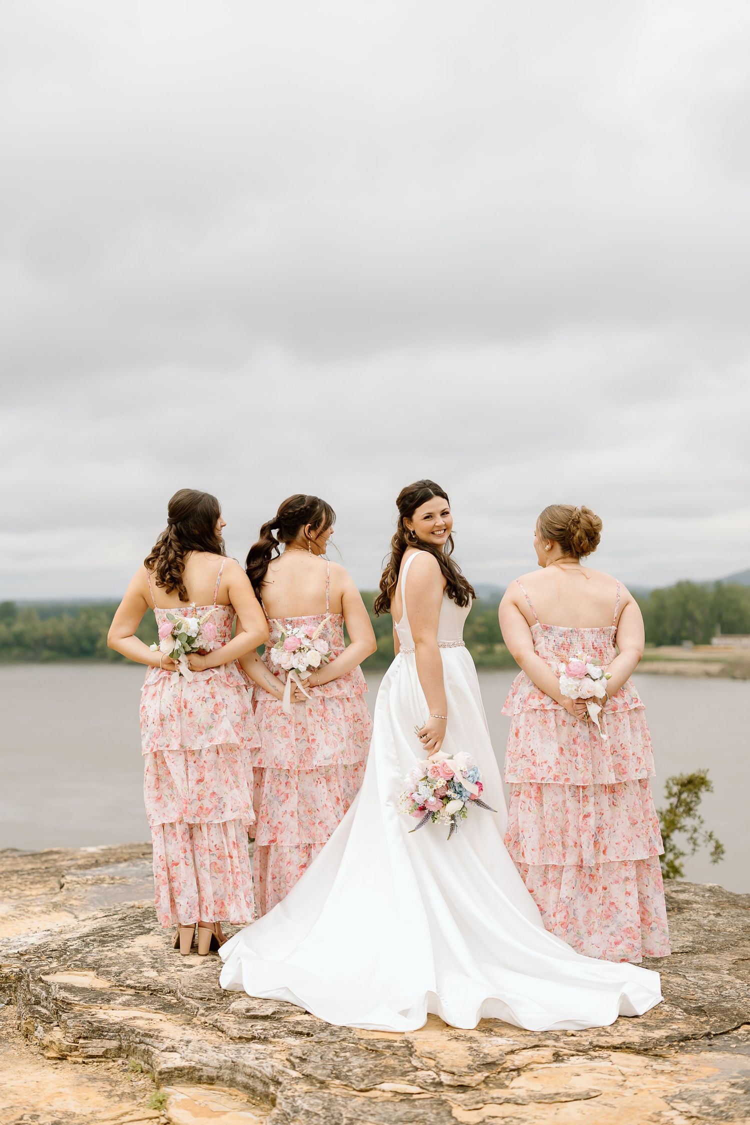 Bridal party in pink floral dresses and white gown pose together on rocky outcrop overlooking scenic lake view.
