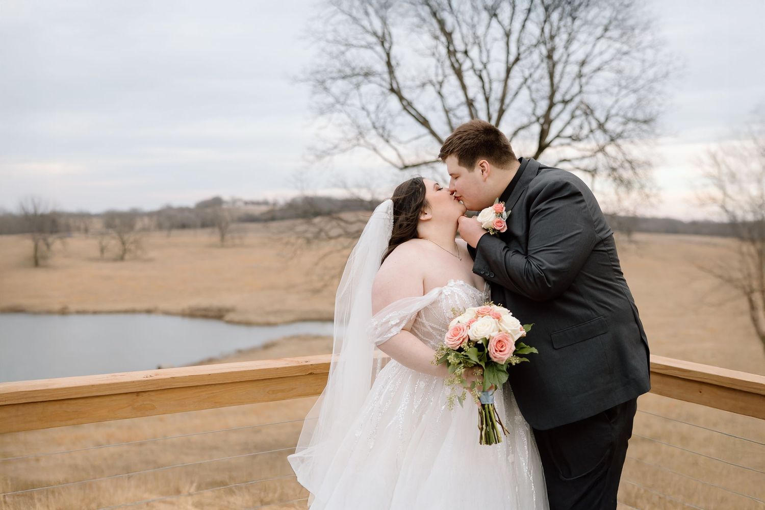 A couple in formal wedding attire embraces in front of a winter landscape with bare trees and open fields.
