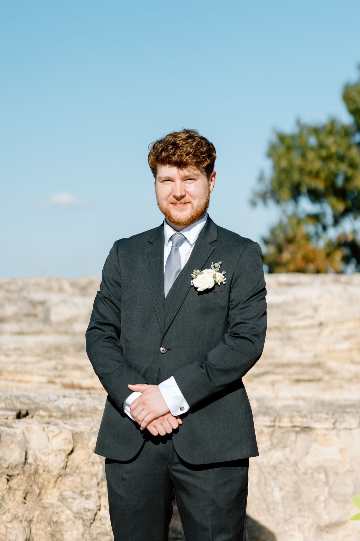 A person in a black suit stands outdoors against a stone wall backdrop on a sunny day.