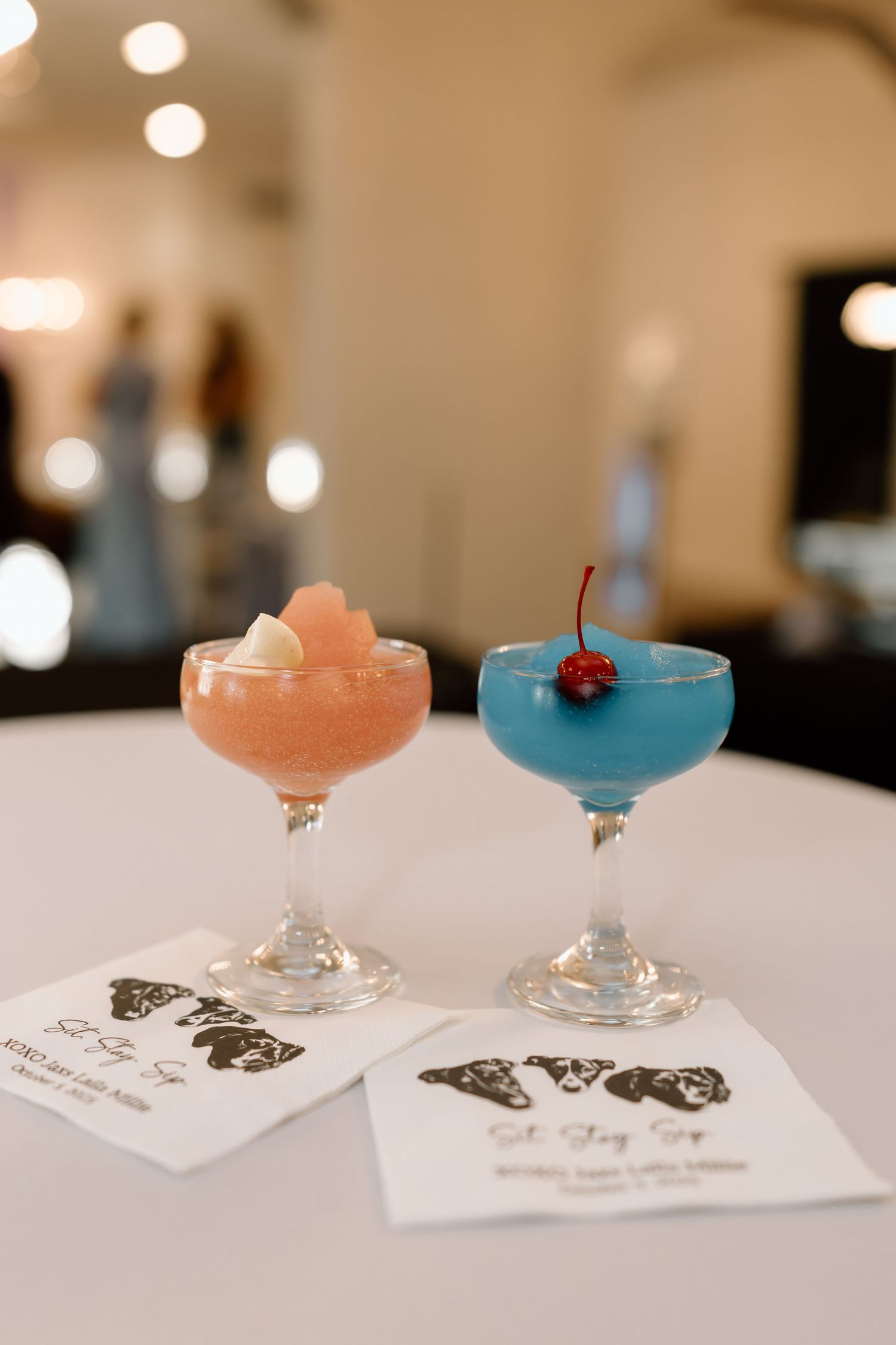 Two elegant cocktails, one pink and one blue, served in coupe glasses with cocktail napkins on a white surface.
