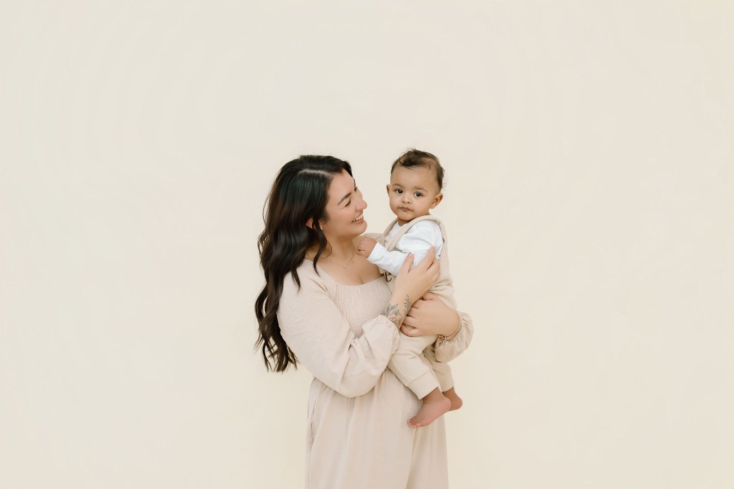 A parent in a cream-colored dress holds a baby against a neutral beige background.