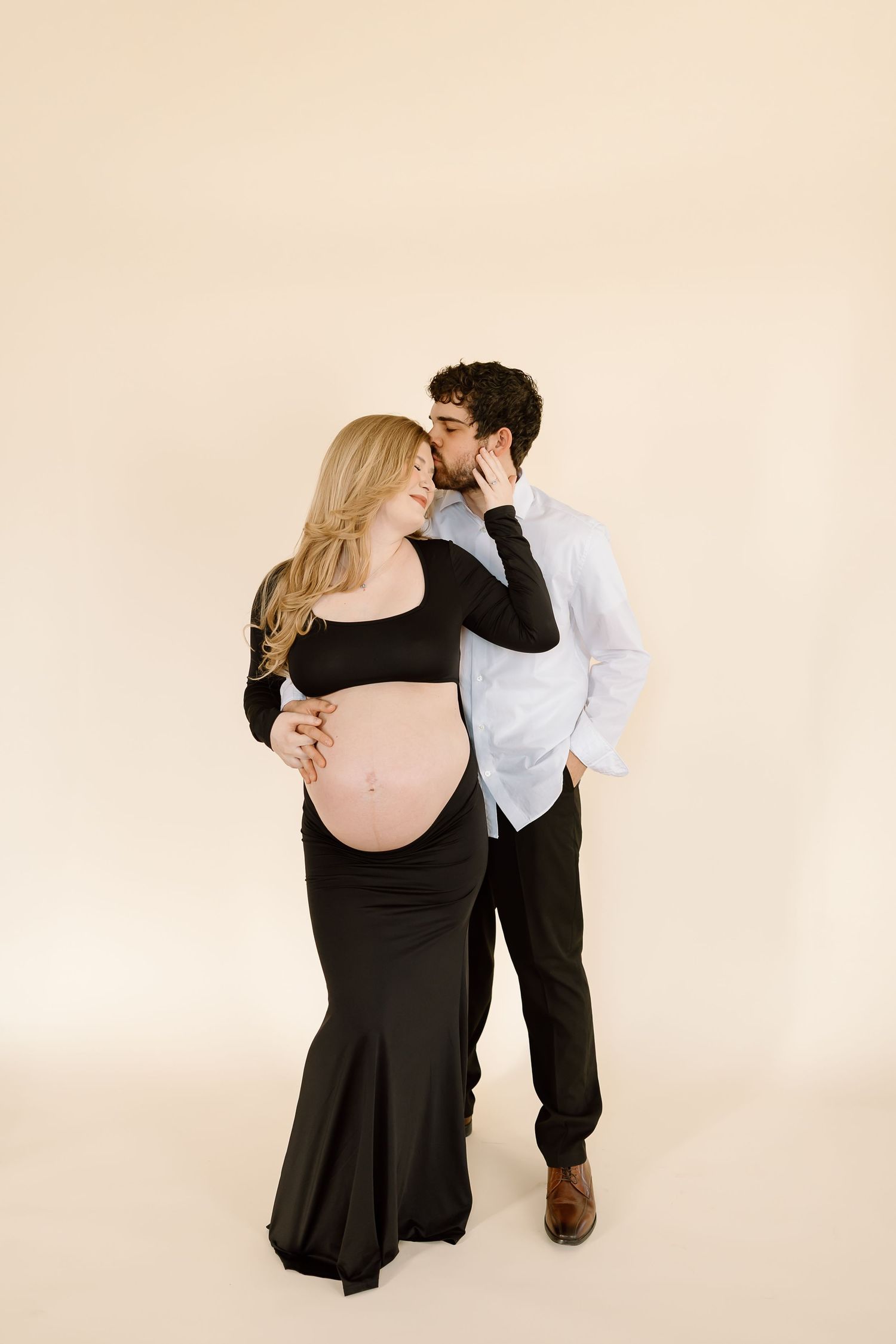 A maternity photoshoot featuring a couple in formal attire posing against a neutral backdrop.