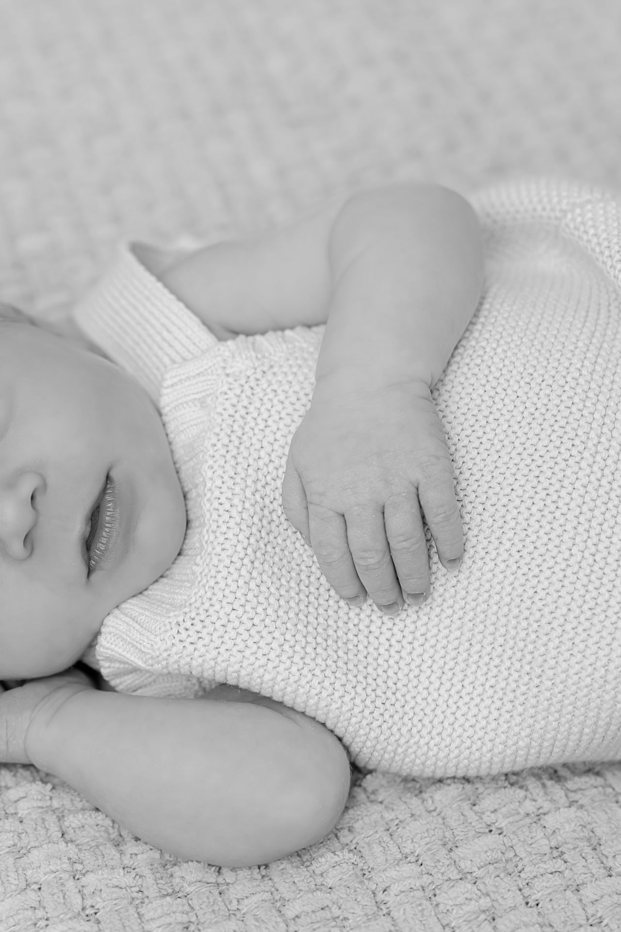 Black and white image of sleeping baby in knit outfit on textured blanket.