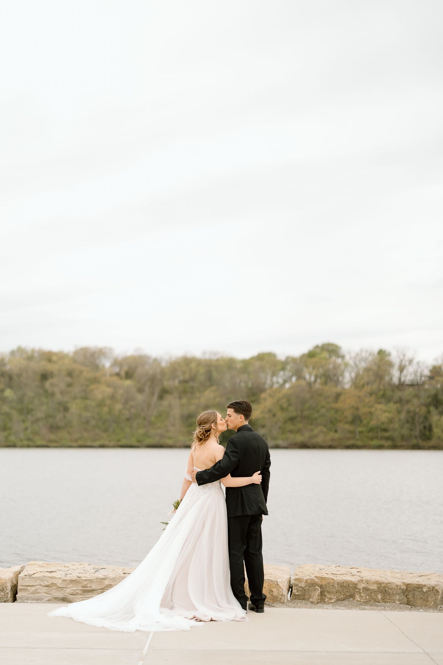 Wedding couple embraces by serene lake with trees in background.