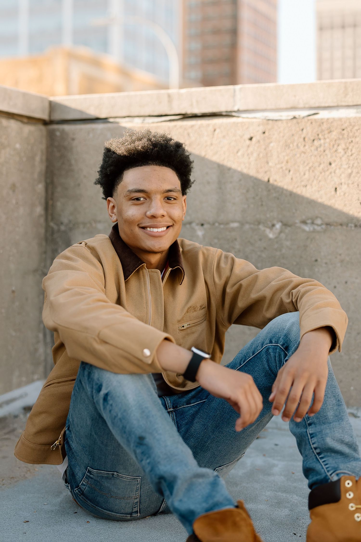 Person in tan jacket sits on concrete floor with urban architecture behind.