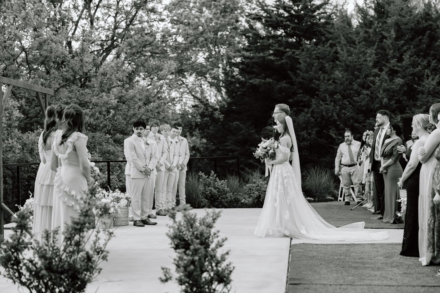 Black and white outdoor wedding ceremony with bride walking down aisle toward groom and wedding party surrounded by lush trees.