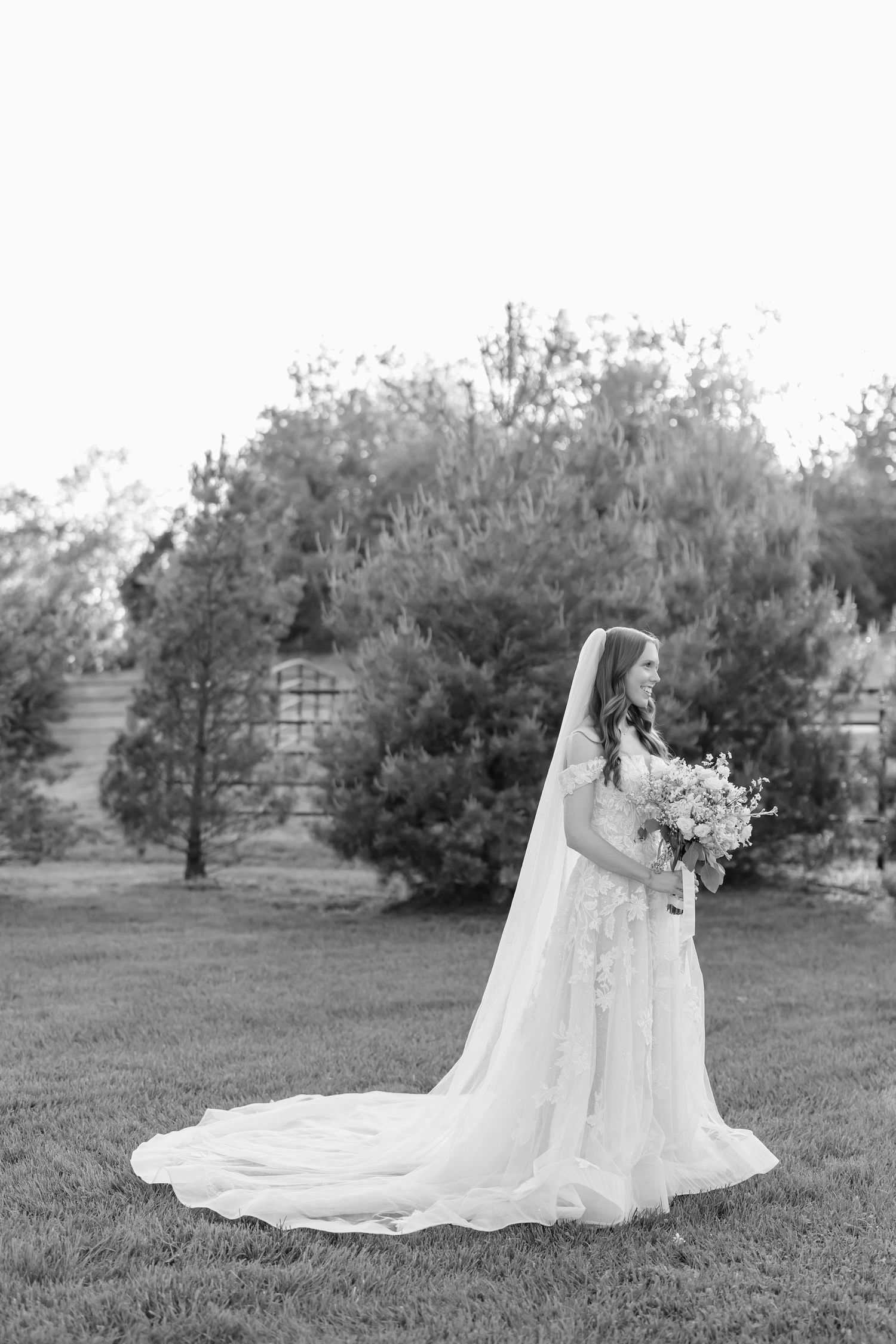 Bride in lace gown and cathedral veil holds bouquet on green lawn with trees and building in black and white photo.
