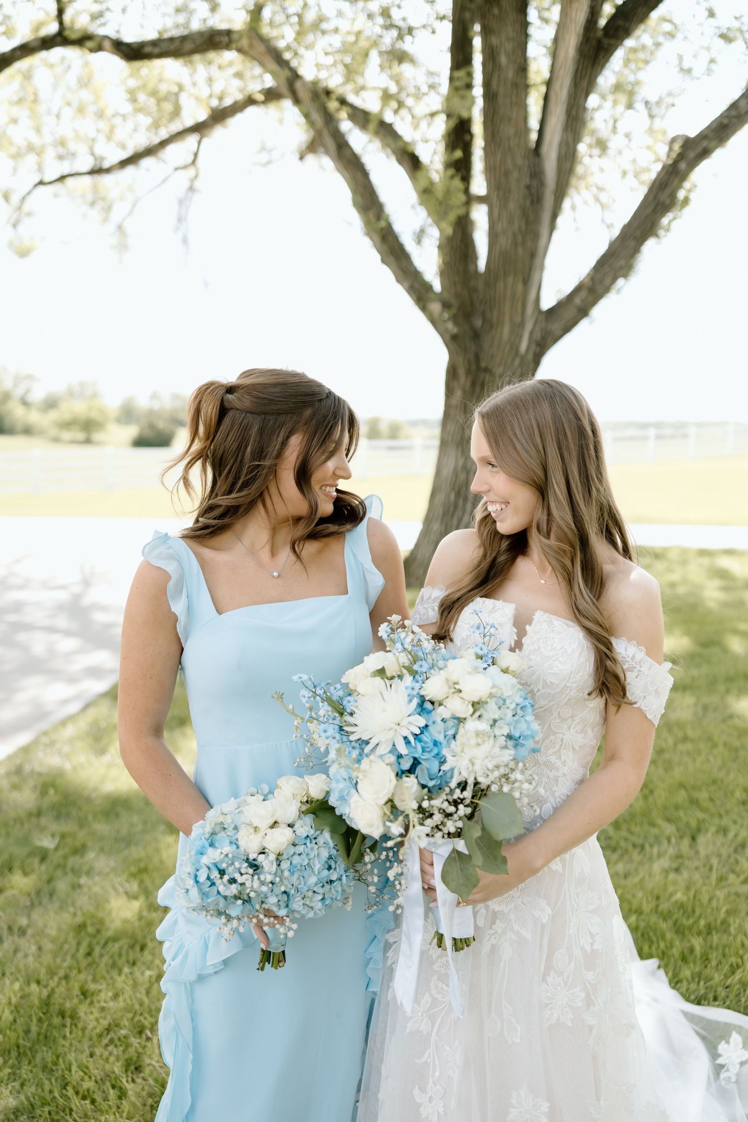 Bride and bridesmaid in light blue dress share a joyful moment holding floral bouquets outdoors.