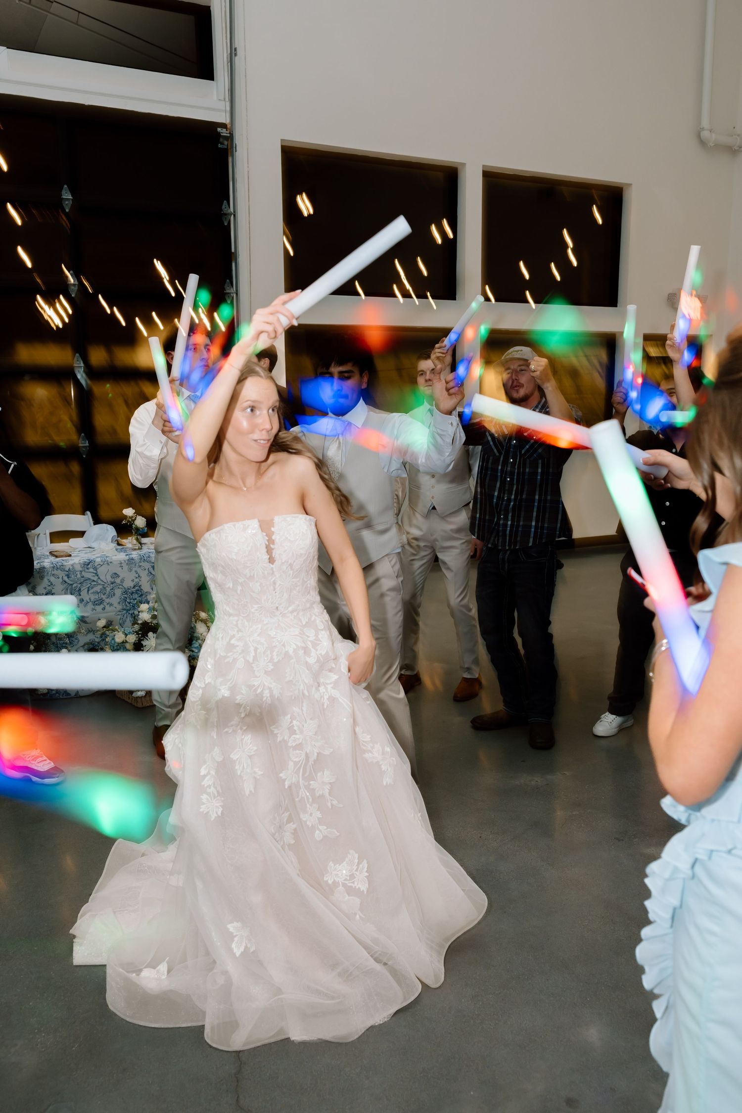 Bride in white lace gown dancing with guests holding colorful LED foam sticks at a wedding reception.