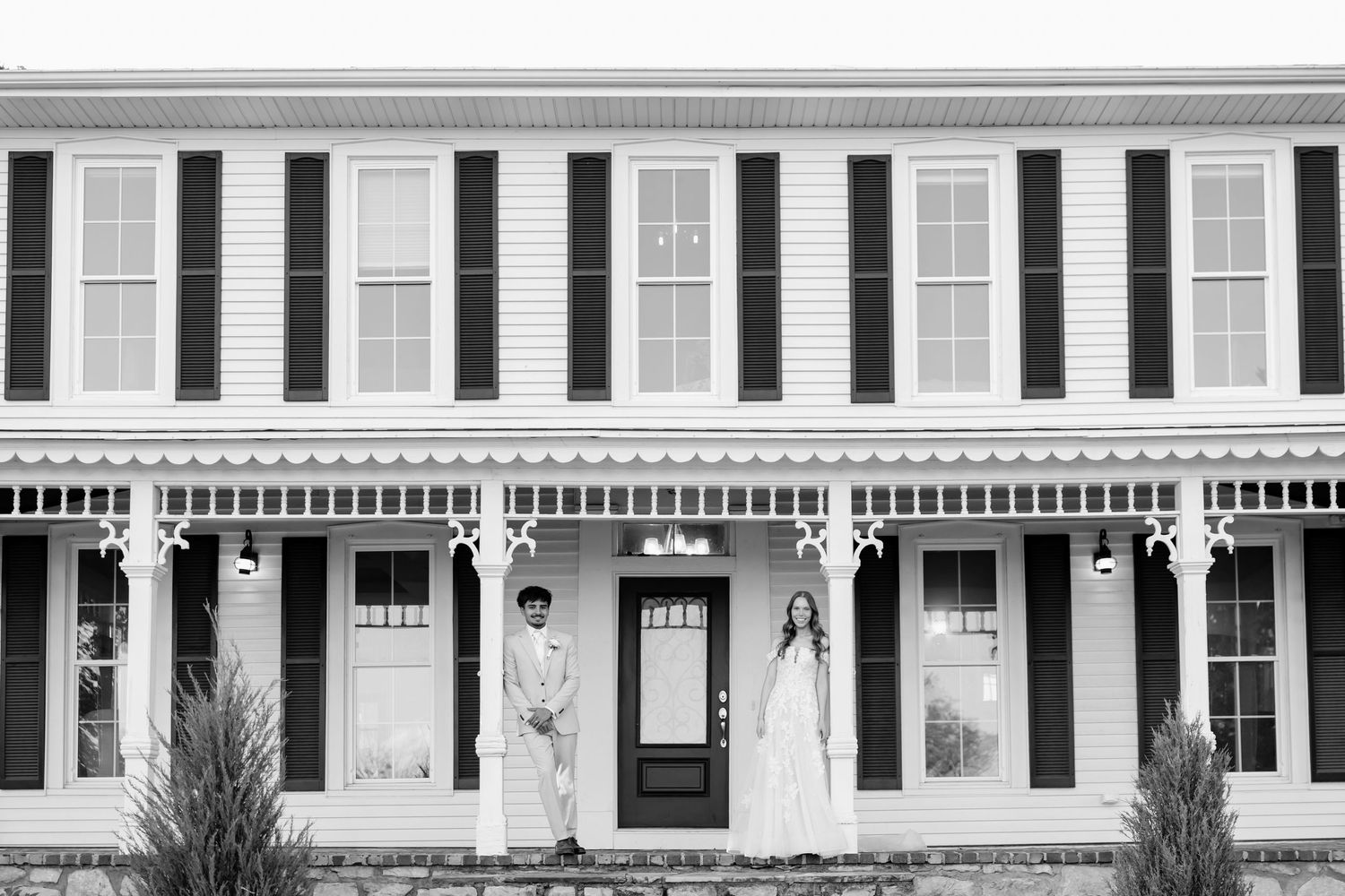Couple stands before a classic white colonial home with black shutters in an elegant black and white wedding portrait.