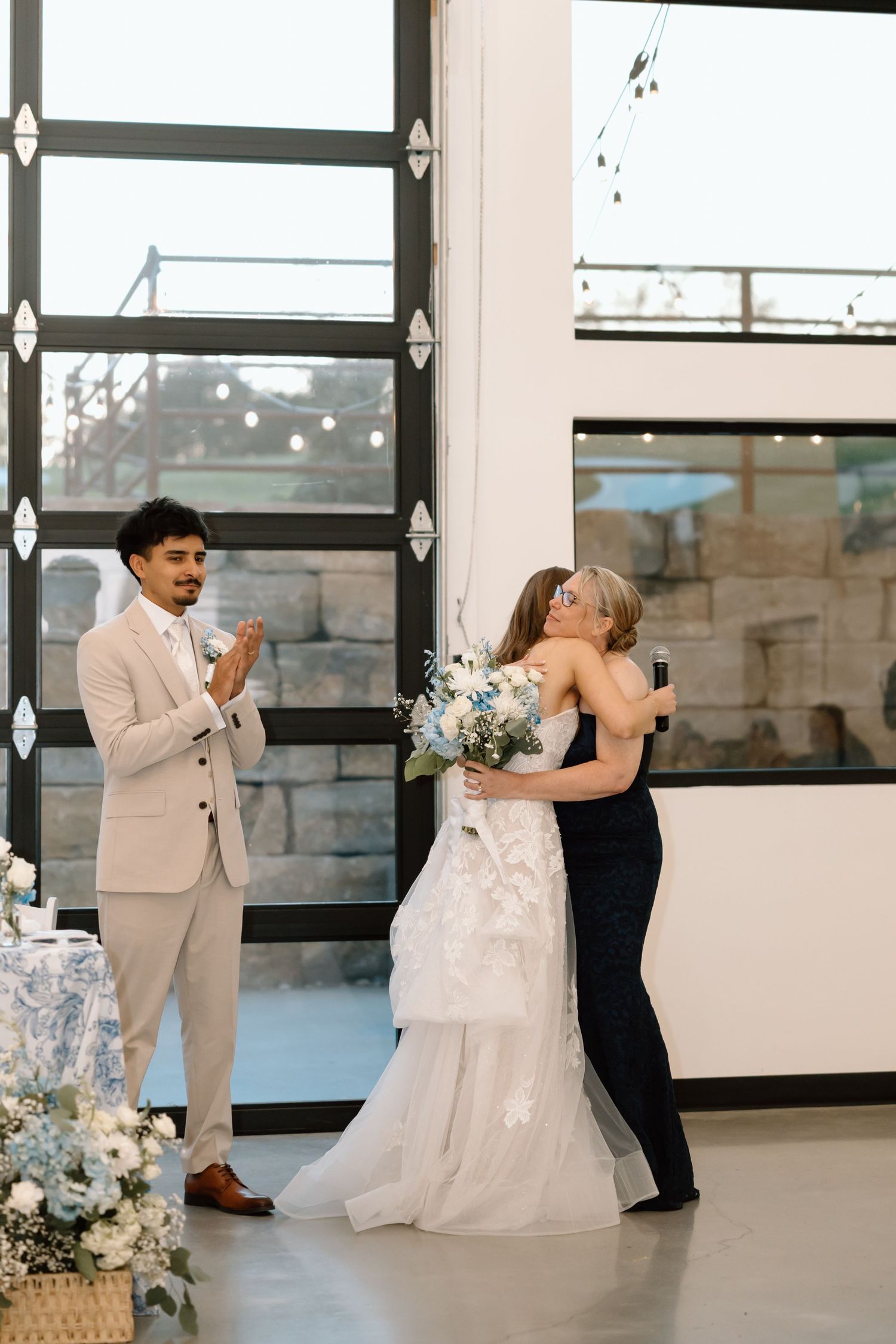 A bride in a white gown hugs a guest while the groom in a beige suit applauds at their wedding reception.