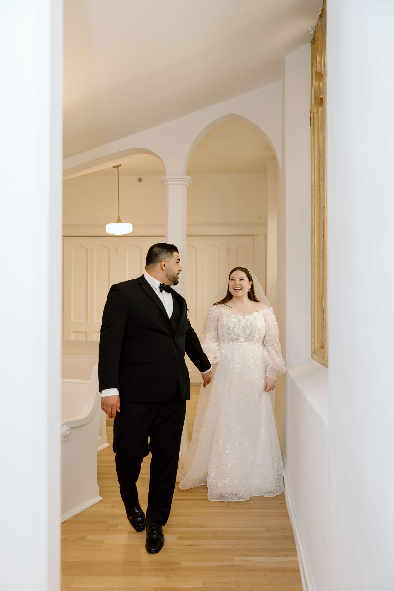 Bride in lace gown and groom in black tuxedo walk hand in hand through elegant white arched hallway.