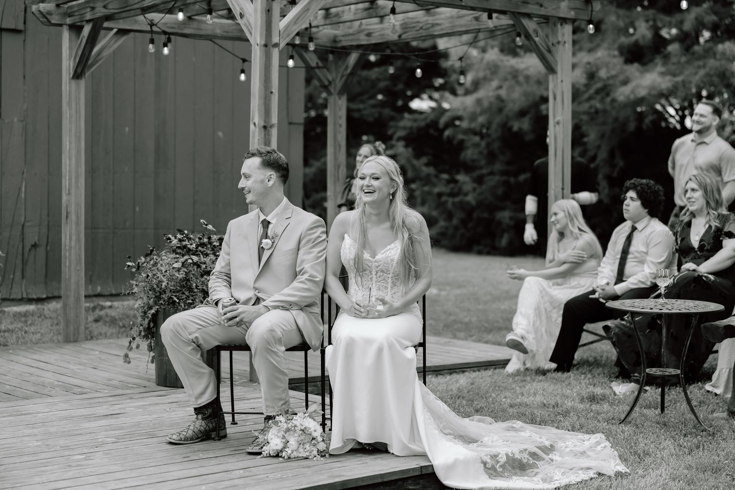 Black and white photo of a smiling bride and groom seated at an outdoor wedding ceremony on a wooden deck.