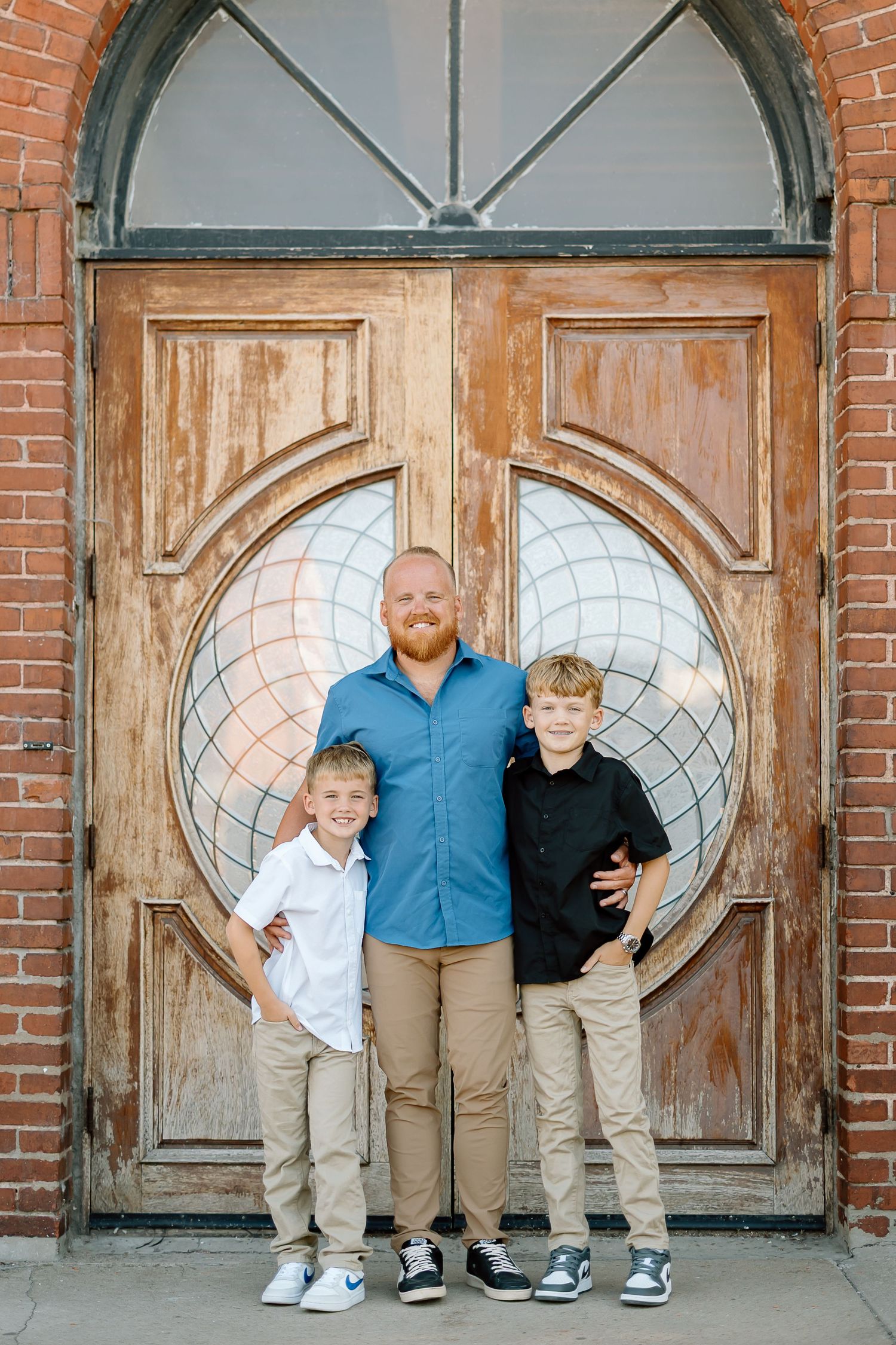 A group stands in front of a rustic wooden door with an oval window design and red brick exterior.