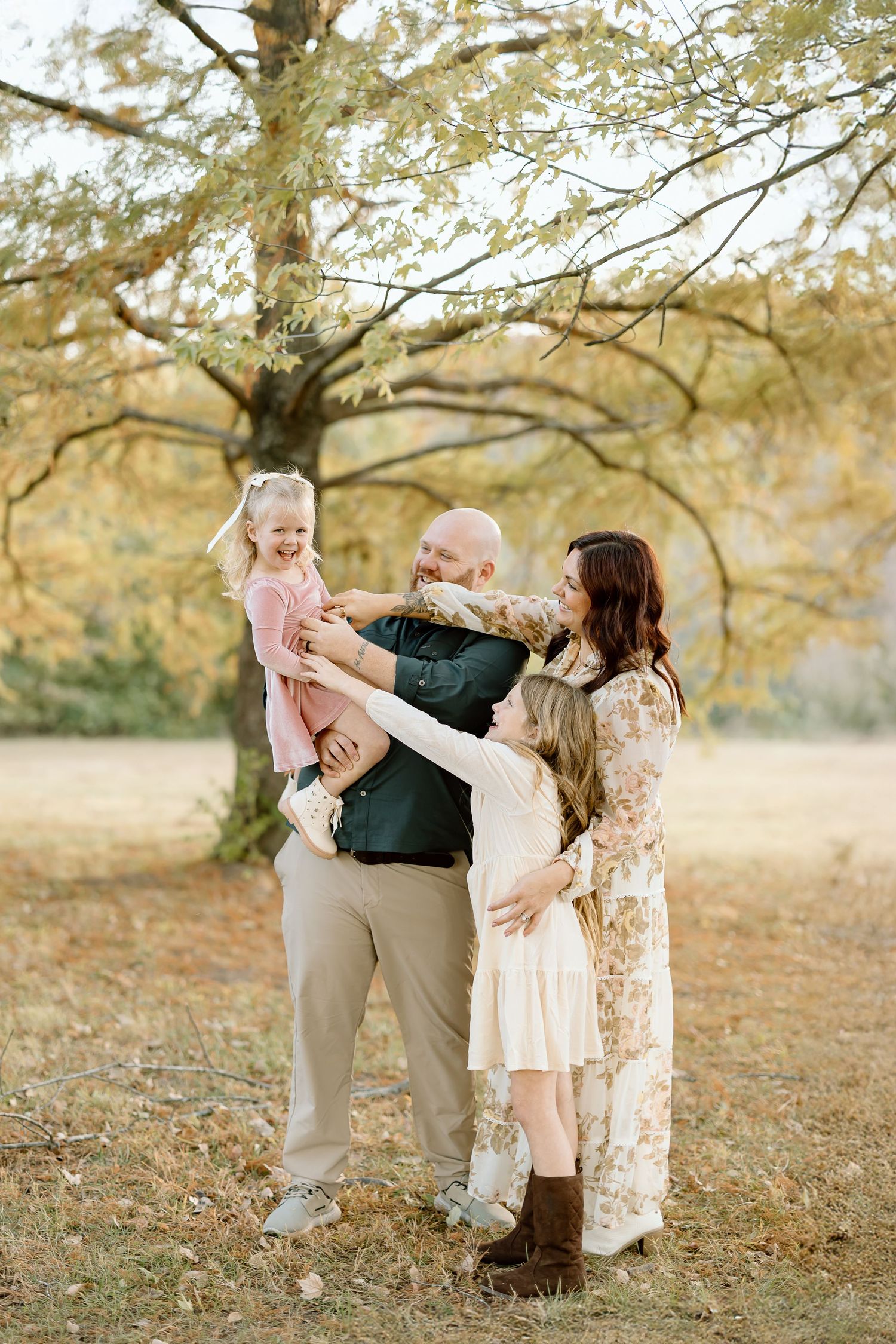 A family shares a playful moment together in a golden autumn field under tree branches at sunset.